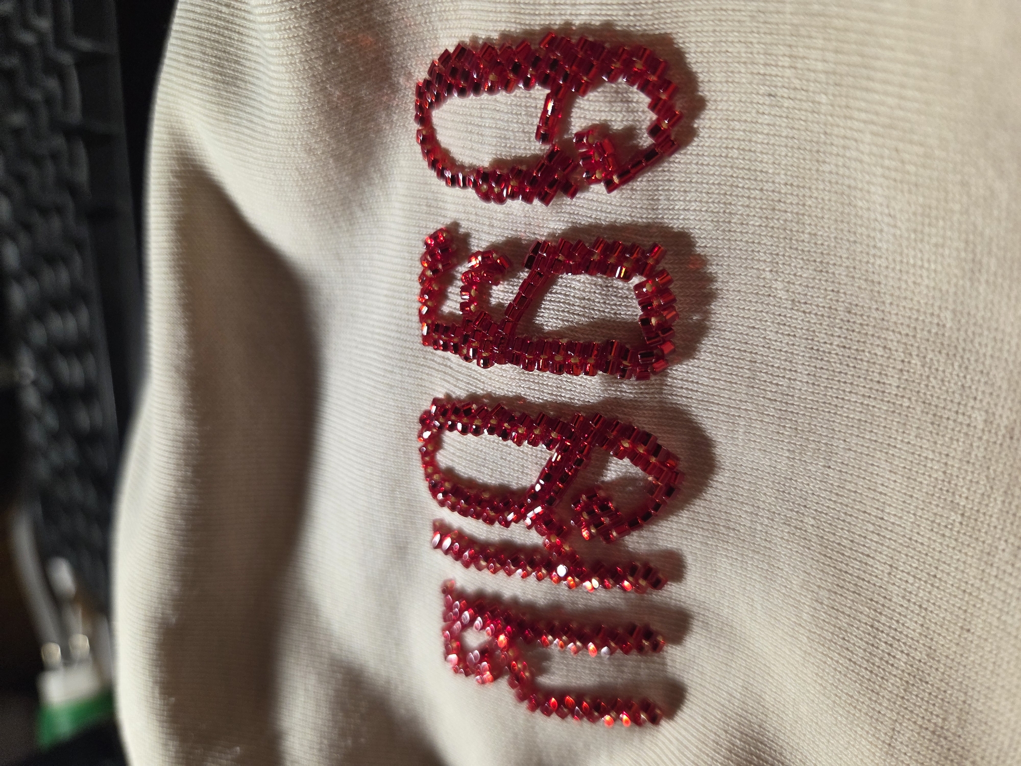 The image shows the number "4969" embroidered with red beads on a light-colored fabric. The fabric appears to be a soft material like cotton or a blend, with a visible knit texture. The beads are small and rectangular, forming the outline of each numeral. The lighting casts shadows from the beads onto the fabric, giving them a slight three-dimensional appearance. The background is blurred, but suggests a dark, possibly electronic device like a keyboard, and some green and white objects. The specific location in the city of Frolovo, Russia cannot be determined from the provided image.