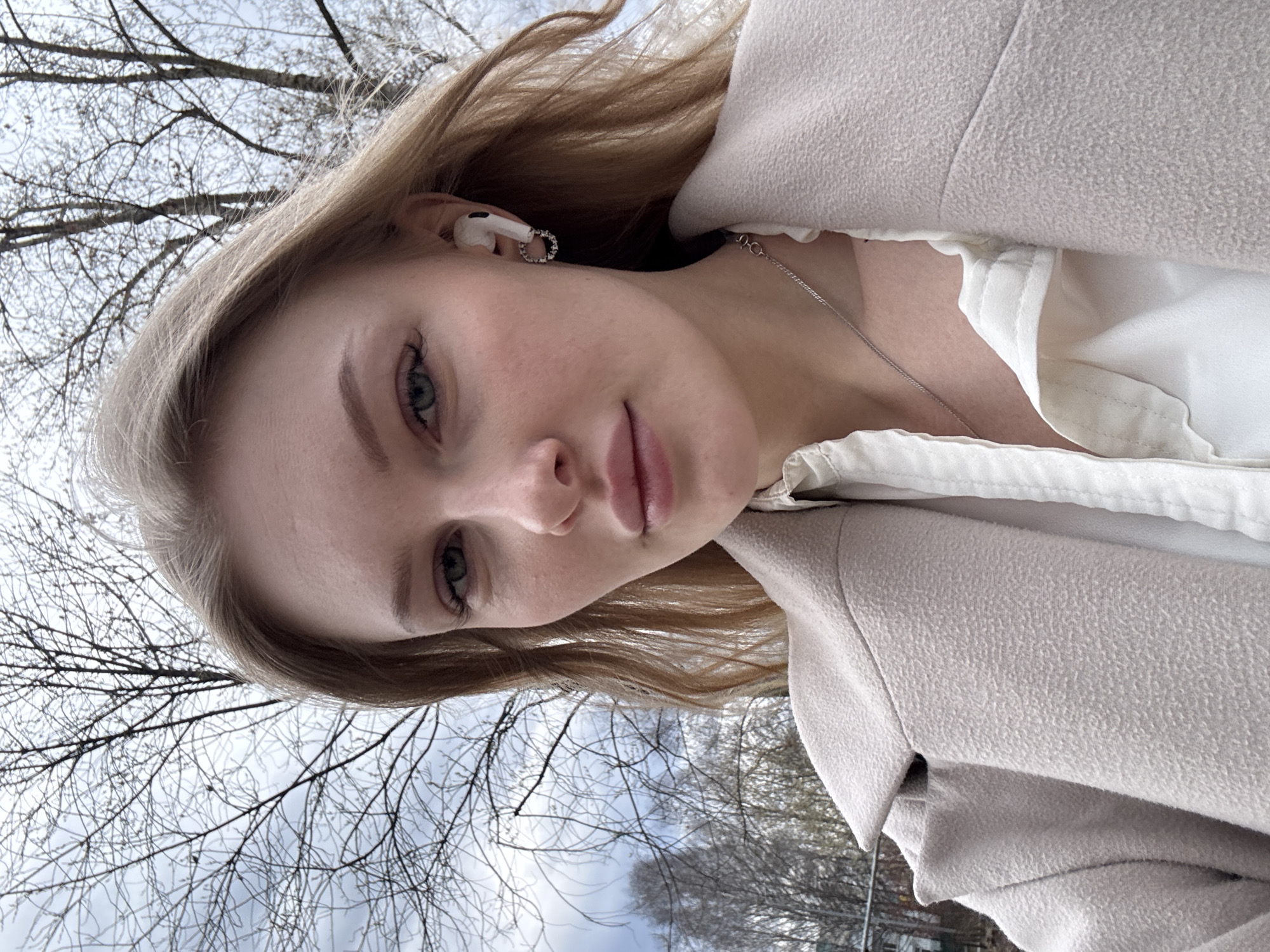 A young Caucasian woman is featured in a close-up outdoor portrait, looking directly at the camera with a calm, neutral expression. She has light blonde hair, fair skin, and light-colored eyes, appearing greenish-blue. Her lips are naturally full with a soft pink hue. In her visible right ear, she wears a white wireless earbud and a small silver hoop earring. A delicate silver chain necklace is visible around her neck.
She is dressed in a white collared shirt, partially seen beneath a warm, light beige or cream-colored coat. The coat has a textured, slightly felted appearance. The photograph is taken from a low angle, looking upwards at the woman and the background.
The setting is outdoors, possibly a park or an urban area in Perm, Russia. The background consists of numerous bare tree branches silhouetted against a bright, overcast sky. The absence of leaves on the deciduous trees indicates a colder season, such as late autumn, winter, or early spring. The overall lighting is soft and diffused due to the overcast conditions. No visible text is present in the image.