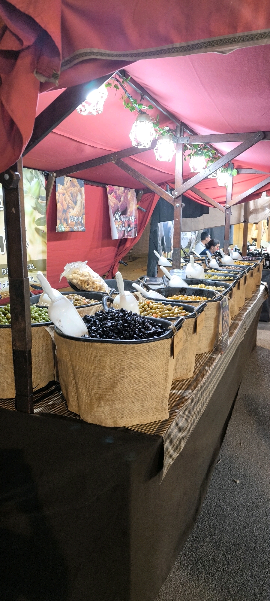 The image shows a market stall in Ripollet, Spain, selling olives and other food items. The stall is covered with a red canopy, and there are hanging lights. In the foreground, there are large tubs filled with various types of olives, including green and black olives. White scoops are placed in each tub for serving. Behind the olives, there are bags of what appear to be dried food items, possibly pork rinds or chips, with labels in Spanish. Two posters with images and text are hanging on the stall's back wall. In the background, two people, a man and a woman, are visible behind the stall, suggesting they are vendors. The stall is set up on a checkered tablecloth, and the ground appears to be asphalt or pavement.