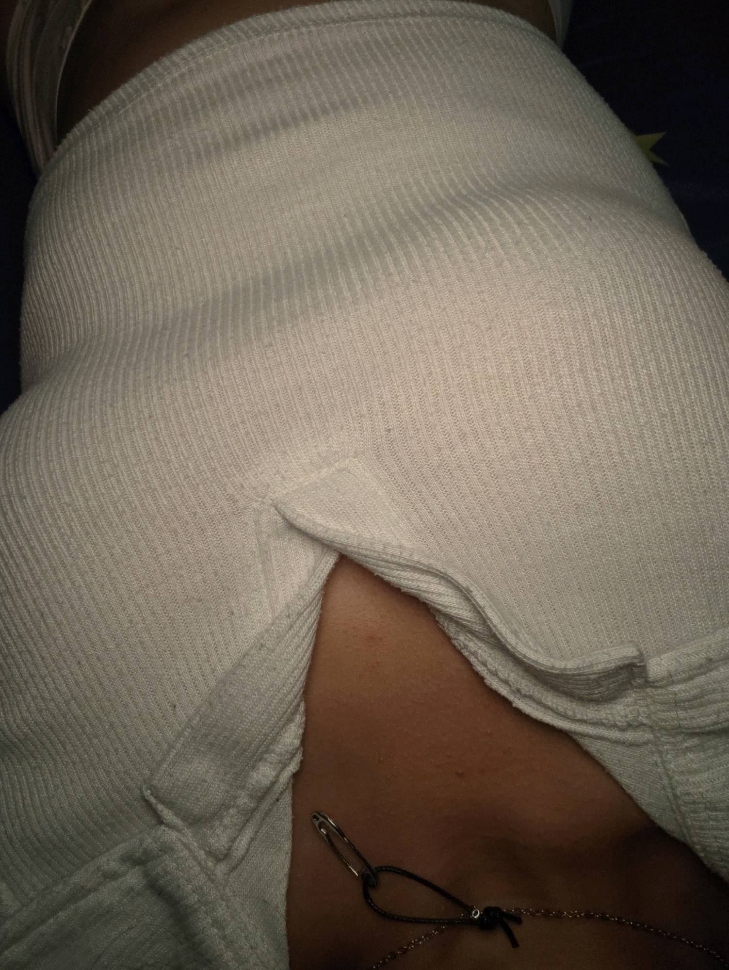 The image shows a close-up view of a person's torso, focusing on the area around the waist and upper hip. The person is wearing a white, ribbed garment that appears to be a tank top or a fitted shirt. There is a slit or opening in the garment, revealing a portion of the person's skin. A safety pin is attached to the garment, and a thin necklace with a dark cord and a metallic chain is visible beneath the opening, resting on the person's neck. The background is dark and indistinct, making it difficult to determine the specific environment. The image provides no information about the location in Pintomakmur, Indonesia.