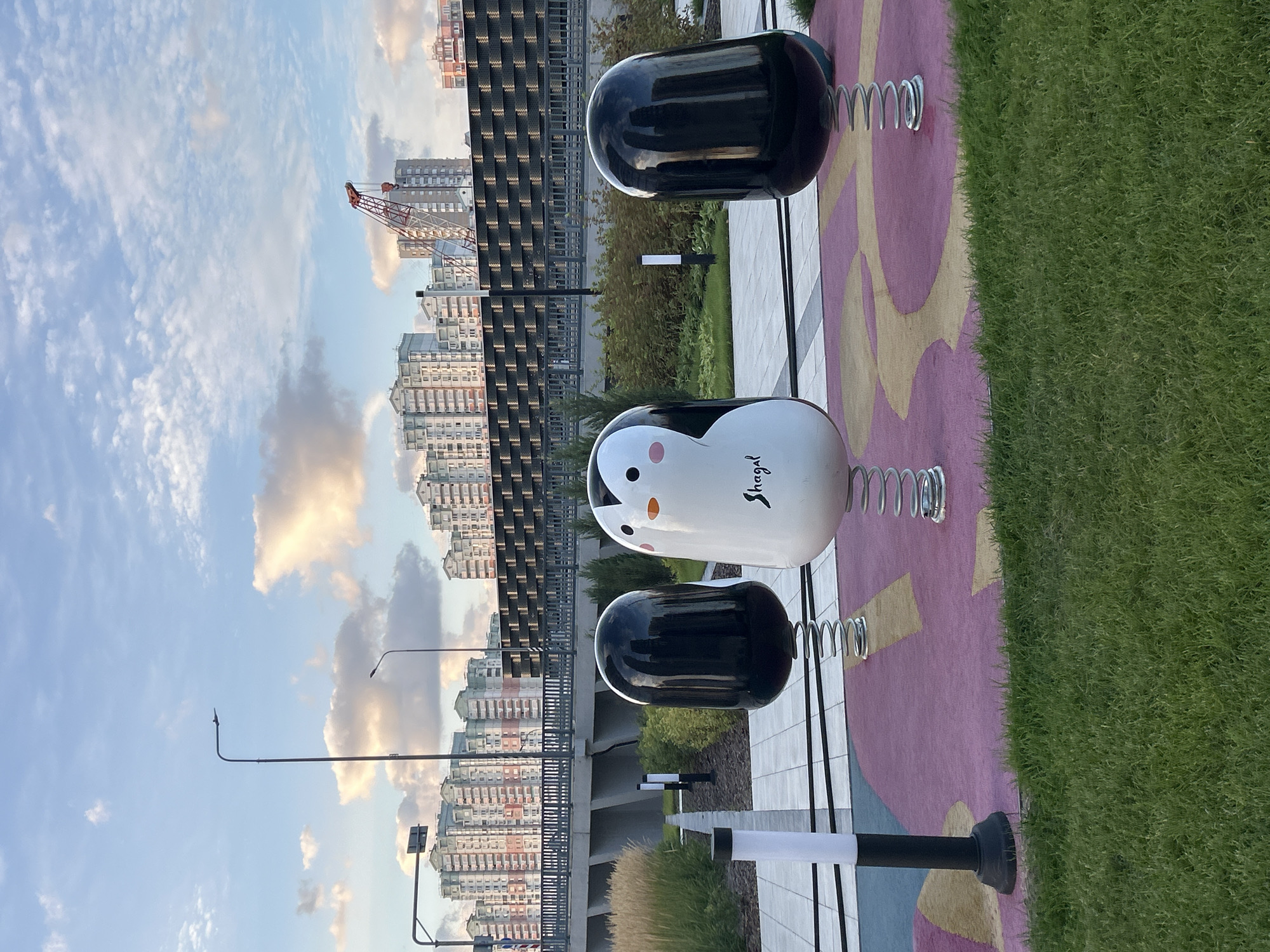 A low-angle outdoor daytime shot captures a playground scene with a sky full of cumulus clouds and a cityscape in the background. In the foreground, three spring-mounted playground figures are visible: two black, oval-shaped figures on either side, and a white penguin in the center. The penguin has black eyes and beak, pink cheeks, and the word "Shagal" written in black script on its chest. The playground figures are set against a pink and yellow abstract patterned ground. Behind the playground equipment, a well-maintained green lawn transitions to a pathway lined with white and black lamp posts. Further back, a dark grey metal fence runs along the edge of an elevated area with manicured bushes and trees. Beyond the fence, a concrete overpass with railings is visible, and in the distance, a dense array of modern apartment buildings with light-colored facades and balconies line the horizon. A construction crane with a red arm stands out against the sky among the buildings. The overall lighting suggests late afternoon or early evening.