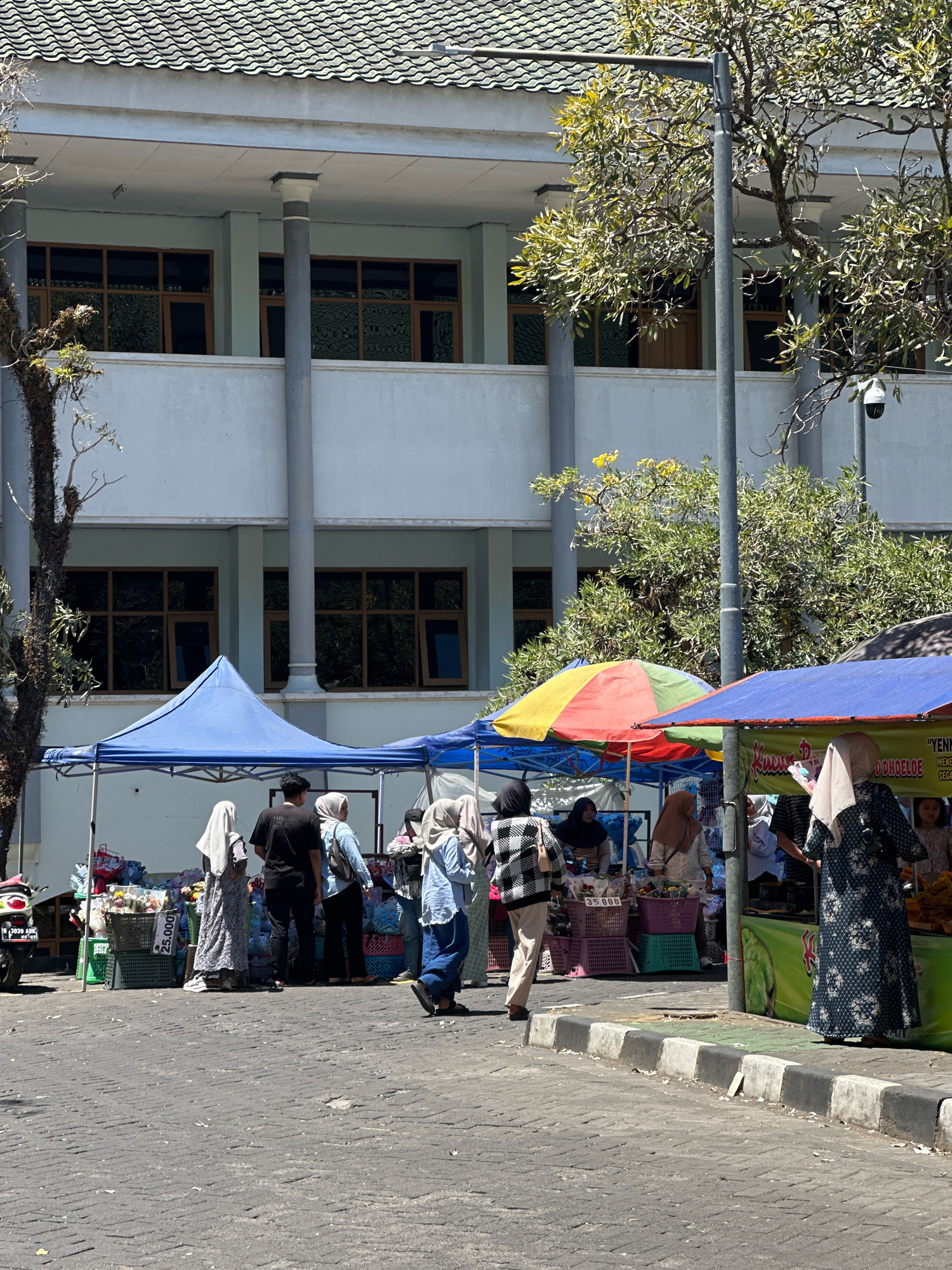 In Malang, Indonesia, a street market unfolds in front of a light blue institutional building with multiple rows of windows. The building's exterior is a pale blue, topped by a dark green tiled roof. The market is set up along a paved street with a distinct curb. Several market stalls, covered by blue, red, yellow, and green awnings, display goods for sale. People in various forms of modest attire, many wearing headscarves, browse and interact at the stalls. One stall features numerous baskets filled with what appear to be produce or merchandise, with a price tag of "25.000". Another stall has a green backdrop with text that is partially visible, including the word "YEN... DDHOELOE". A motorcycle is parked to the left of the scene. Trees with green foliage, some bare branches, and a street lamp are also visible, casting shadows on the market area. The sky is clear and bright, indicating a sunny day.