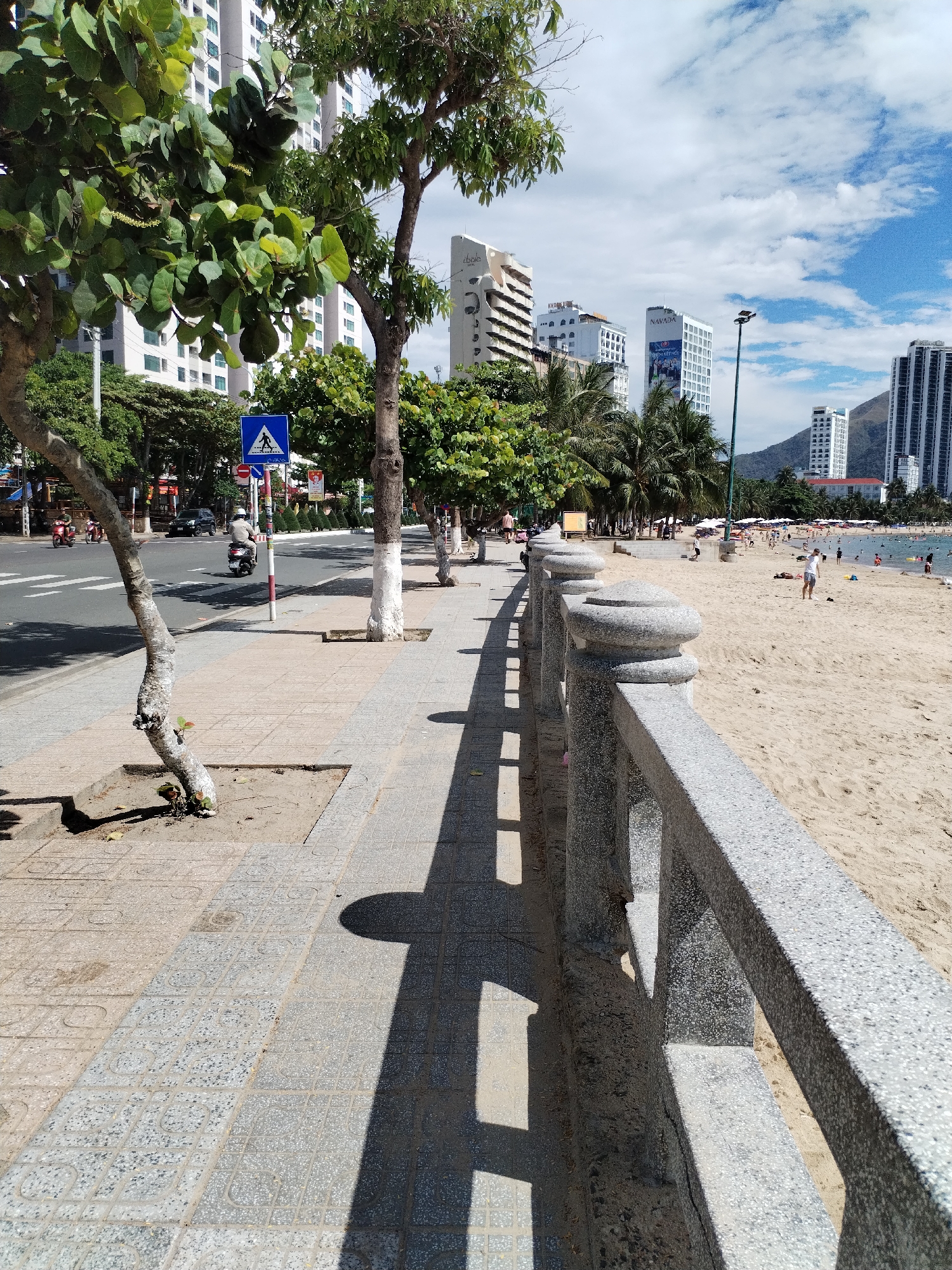 This media file captures a vibrant, sunny street scene along a beach in Bac Nha Trang Ward, Vietnam, likely during midday or early afternoon given the strong shadows.
In the foreground, a textured grey granite-like balustrade runs parallel to the viewer, separating a tiled sidewalk from a sandy beach. The sidewalk is paved with light grey tiles featuring a repeating circular pattern. To the left of the balustrade, tall trees with green foliage cast prominent shadows across the sidewalk. Some tree trunks are painted white.
To the left of the sidewalk is a paved road with multiple lanes, where several motorbikes, including one with a rider wearing a helmet, are visible. A blue square sign indicating a pedestrian crossing stands beside the road. In the distance along the road, more trees and buildings can be seen.
On the right, beyond the balustrade, a wide sandy beach is bustling with people. Many individuals are swimming in the clear water or relaxing on the sand, some under beach umbrellas.
In the background, modern, multi-story buildings, characteristic of urban coastal development, line the shore. One prominent white building displays the word "NAVADA," and another building has a partially visible sign that appears to read "Cholo" or "Ebola." Distant mountains frame the cityscape. The sky above is blue with scattered white, fluffy clouds.