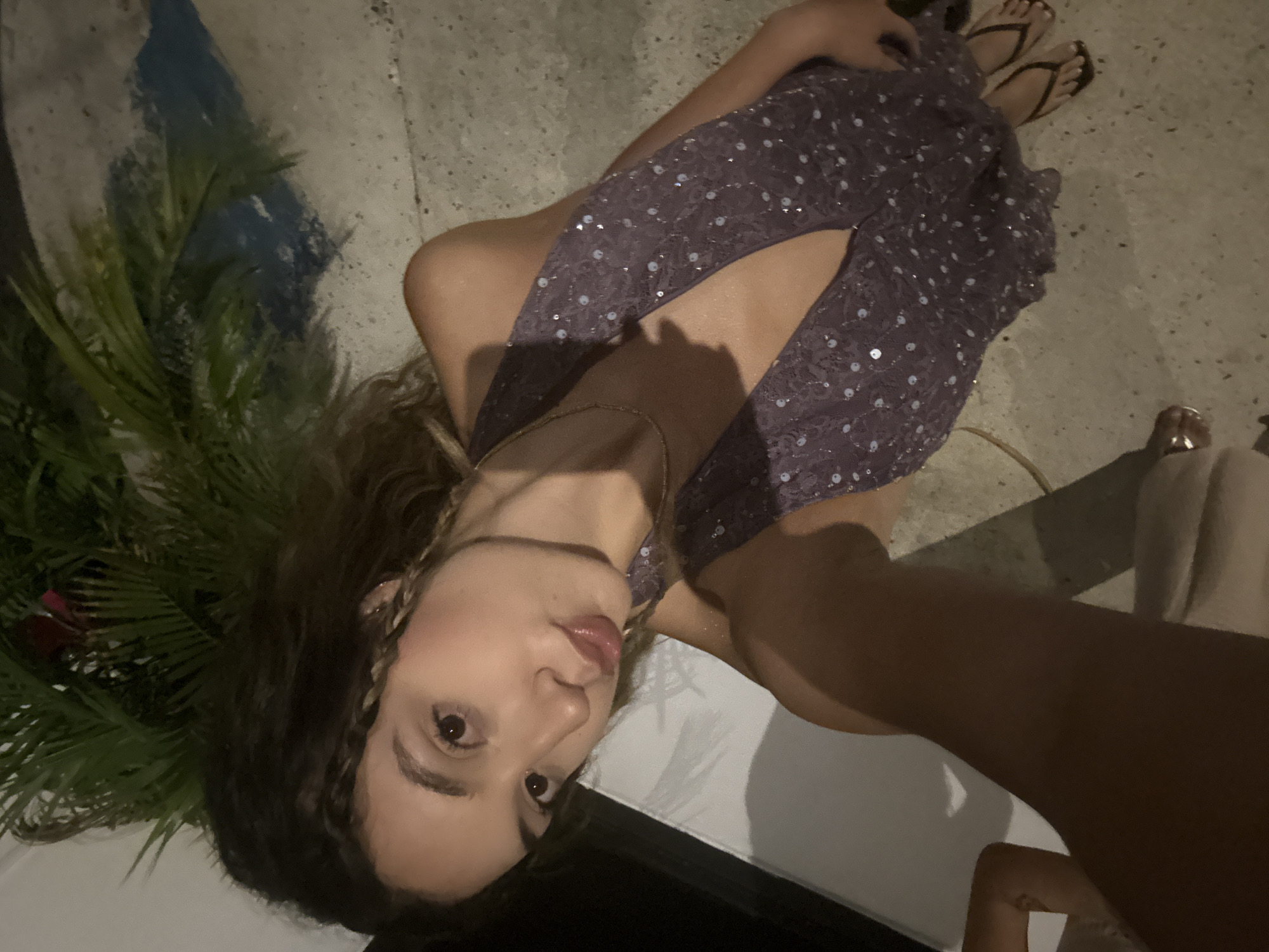 A young woman with curly brown hair is taking a selfie outdoors at night. She is wearing a sleeveless, dark purple, sparkly dress and flip-flops. The background shows a concrete walkway, some greenery, and a white wall. The location is San Juan, United States of America.