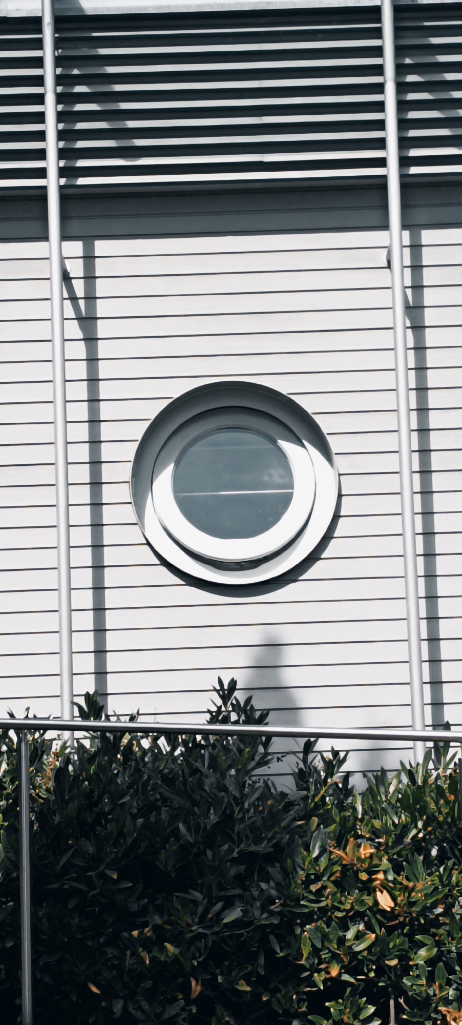 The image shows a close-up of a modern building exterior, likely in the city of Fellbach, Germany, given the context. The facade is composed of horizontal white slats and features a prominent, circular, porthole-style window with a silver frame. To the left of the window, two vertical silver poles run up the building. A shadow of a pole is cast on the wall to the left of the window. In the foreground, a dark green hedge with some brown leaves is visible, partially obscured by a silver railing. The lighting suggests it is daytime, with sunlight creating shadows on the building's surface. No people are visible in the image.