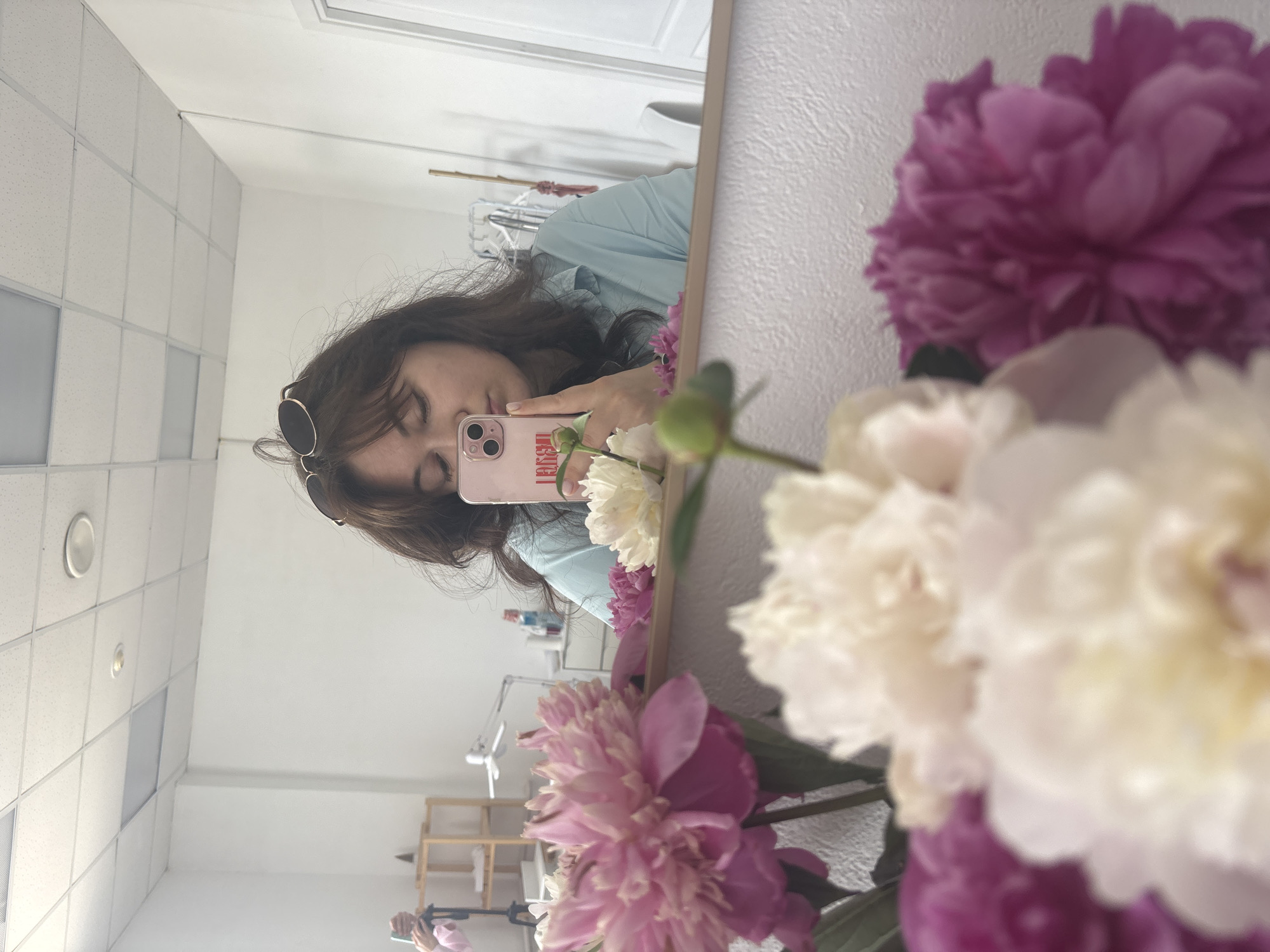 In this image, a woman with dark brown hair and bangs is captured in a mirror selfie, holding a pink smartphone. She is wearing sunglasses on top of her head and a light blue collared shirt. Her eyes are closed, and she is leaning in towards the phone. In the foreground, a bouquet of peonies is visible, with large pink and white blooms. The background shows a room with white walls, a tiled ceiling with lights, and a closet with clothes on hangers. A shelf unit with items on it is also visible to the left. The reflection in the mirror indicates that the photographer is likely in a room, possibly a home or a studio, located in the city of Chaikovsky, Russia. The overall impression is a casual, personal moment captured in a domestic or creative space.