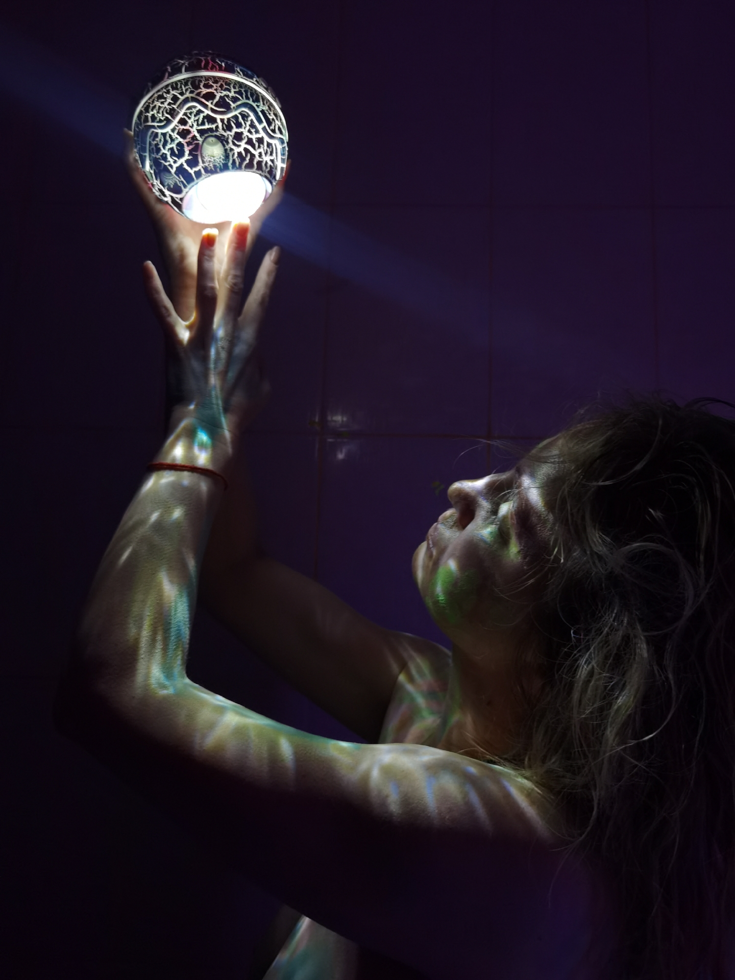 This image features a person, likely a woman, seen from the mid-torso up, captured in profile against a dark, possibly tiled, purple background. Her head is tilted slightly upwards, and her eyes gaze in the same direction. Her light-colored, wavy hair frames her face and flows down her back.
Her hands are raised above her head, reaching for and seemingly holding a glowing, spherical object. The sphere is intricately designed with a white or silver, crackled or organic pattern over a darker surface, and emits a bright, intense white light from its base. This light forms a visible beam upwards and casts vibrant, multi-colored patterns (predominantly greens, blues, and yellow-greens) onto the person's arms, chest, and face, suggesting the patterns from the sphere are projected onto her skin. A thin red bracelet is visible on her left wrist. Her fingernails are painted a reddish hue.
The activity depicts a focused interaction with the luminous sphere, possibly evoking a sense of wonder, concentration, or spiritual connection. The overall setting is dark, emphasizing the glow of the sphere and the projected light patterns, suggesting it is either night or a dimly lit indoor space. There is no visible text in the image.