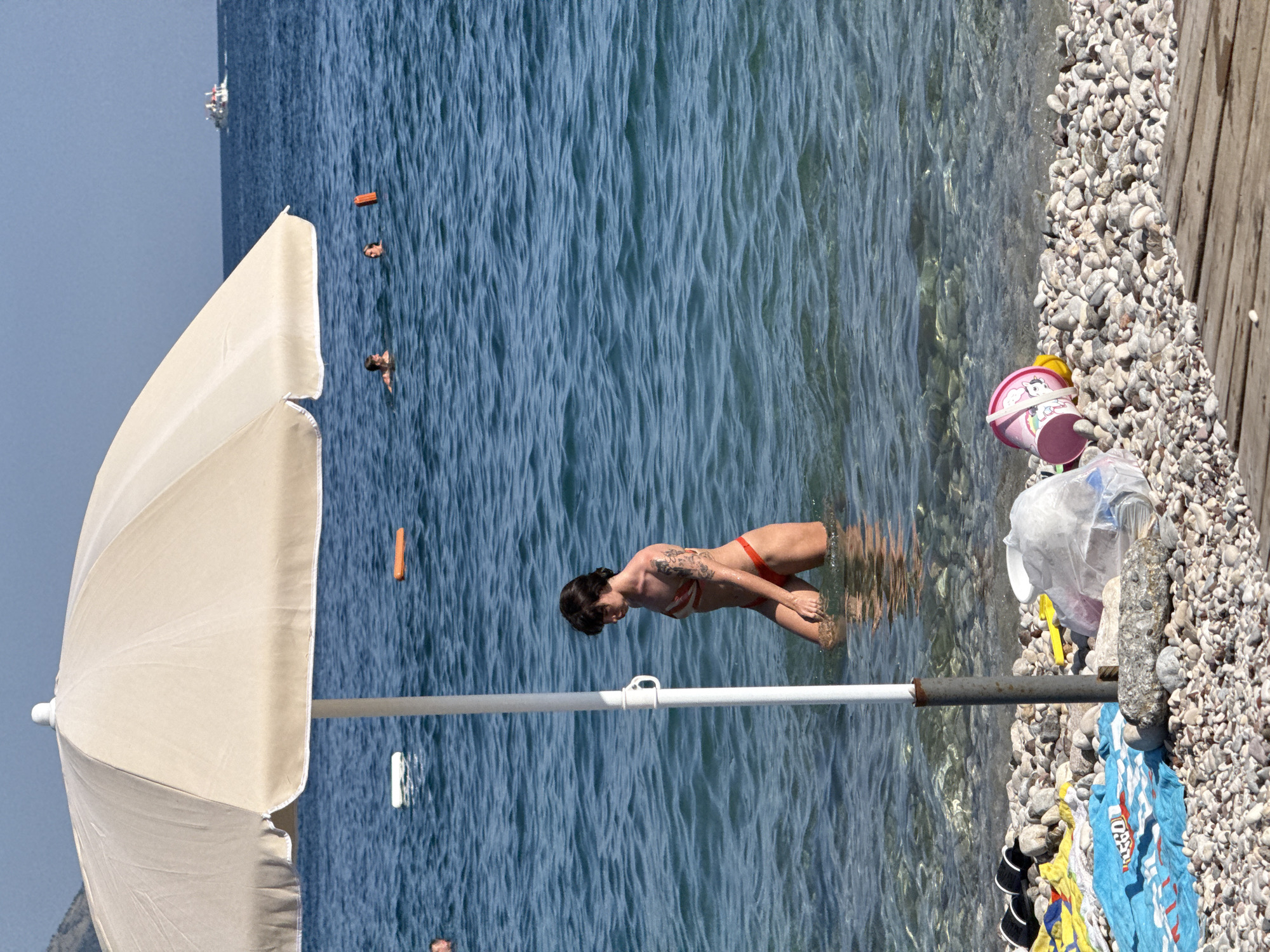The image depicts a sunny day at a pebble beach in Budva, Montenegro. A large, light-colored umbrella stands in the shallow, clear blue water, its pole secured to the beach. A young woman, wearing a red bikini and with visible tattoos on her arms, is wading in the water, looking down. In the background, the sea stretches out, with a few white buoys and an orange float visible. Further out, a boat is visible on the horizon. On the shore, near the umbrella's base, are various beach items: a blue towel with text, a yellow towel, a pair of black flip-flops, a pink bucket with a unicorn design, a yellow toy spade, and a clear plastic bag filled with debris. The beach itself is covered in smooth, grey and white pebbles.