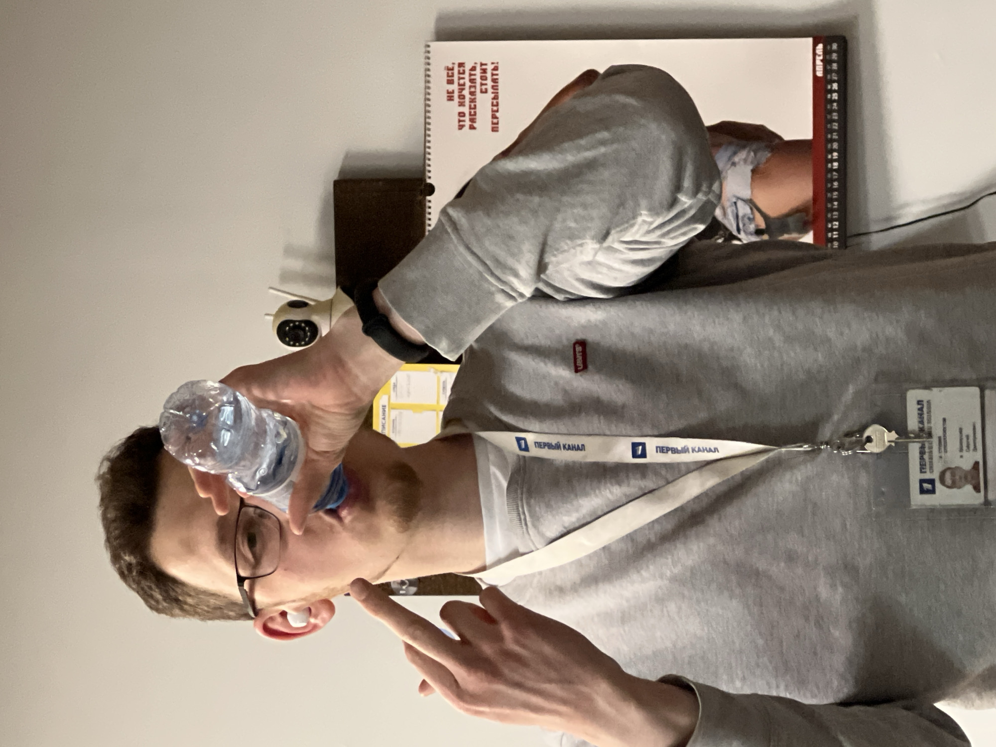 An indoor, vertically oriented photograph, rotated 90 degrees clockwise, captures a man drinking water. The man, who appears to be in his 20s or 30s, has short dark hair, glasses, and a light beard. He is wearing a grey sweatshirt with a small red "Levi's" tag visible on the left side of his chest, over a white t-shirt. A white lanyard with "ПЕРВЫЙ КАНАЛ" (Channel One) printed repeatedly hangs around his neck, holding an ID badge with his photo and "ПЕРВЫЙ КАНАЛ" also visible. A smart band is on his left wrist.
He is seen drinking from a small plastic water bottle, tilting his head back to do so, with his right index finger slightly raised. The background is a plain light-colored wall. On a surface behind him, a small white security camera is visible to the left of his head, and a dark brown object, possibly a wallet or book.
To the right of the man's head, resting against the wall, is a white spiral-bound notebook or calendar. Clearly legible red text on its cover reads: "НЕ ВСЁ, ЧТО ХОЧЕТСЯ РАССКАЗАТЬ, СТОИТ ПЕРЕСЫЛАТЬ!" which translates to "NOT EVERYTHING YOU WANT TO TELL IS WORTH SENDING/FORWARDING!" The word "АПРЕЛЬ" (April) and a numerical scale (1-30, with "28 29 30" visible) are also on the notebook. Partially visible yellow text to the far left reads "ПИСАНИЕ" (Writing/Scripture).
The man's neutral expression is focused on his activity. The presence of the "Channel One" lanyard and ID, along with the visible Russian text, suggests a professional setting, possibly related to media in Khimki, Russia.