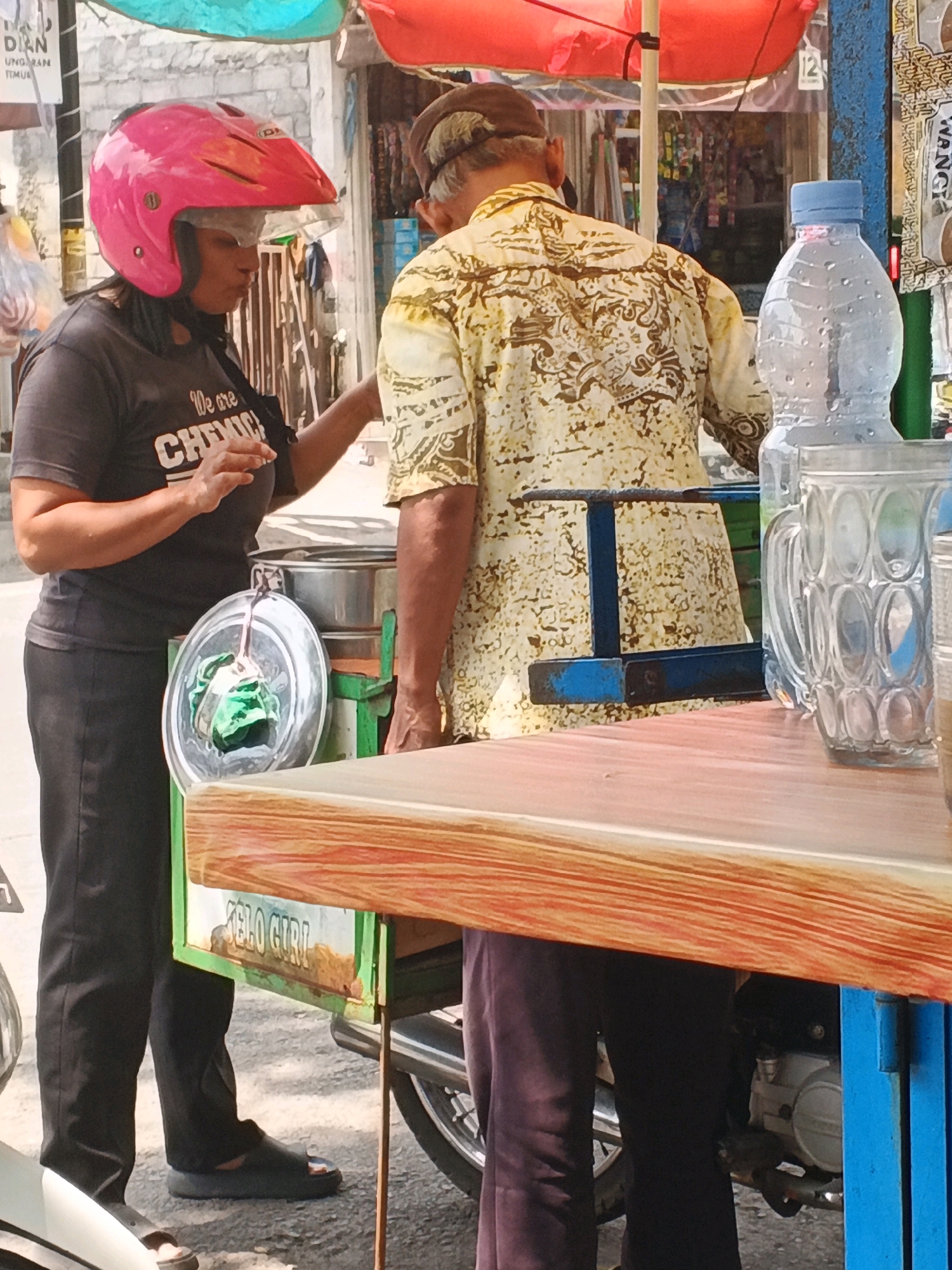 In Kalirejo, Indonesia, a street vendor wearing a pink helmet stands beside a cart, interacting with a customer. The vendor, dressed in a black t-shirt and pants, is in profile, facing the customer. The customer, wearing a patterned yellow shirt and dark pants, is seen from the back. The cart has a green frame and a wooden top, with signage that reads "ELO GIRL". A stainless steel container and a clear plastic bowl with green contents are on the cart. A bottle of water and a glass mug are on the wooden surface. The background shows a market or roadside setting with stalls and a red awning overhead. The scene is illuminated by natural daylight.