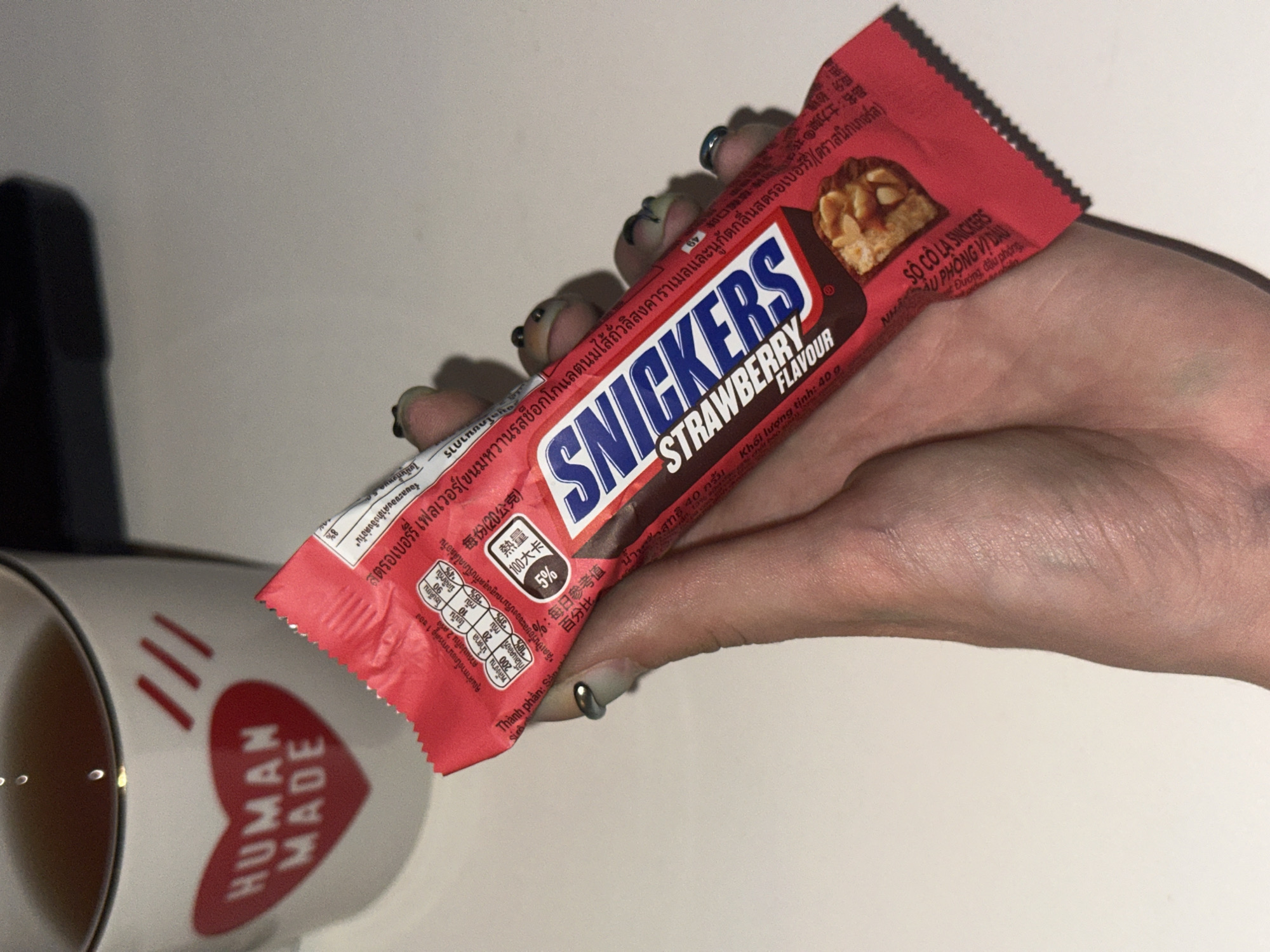 A hand holds a Snickers Strawberry flavor candy bar. The candy bar is red with blue lettering that says "SNICKERS STRAWBERRY FLAVOUR". The background shows a white mug with a red heart and the words "HUMAN MADE" on it. There is also a cup of dark liquid. The scene appears to be indoors on a white surface. The image was taken in Moscow, Russia.