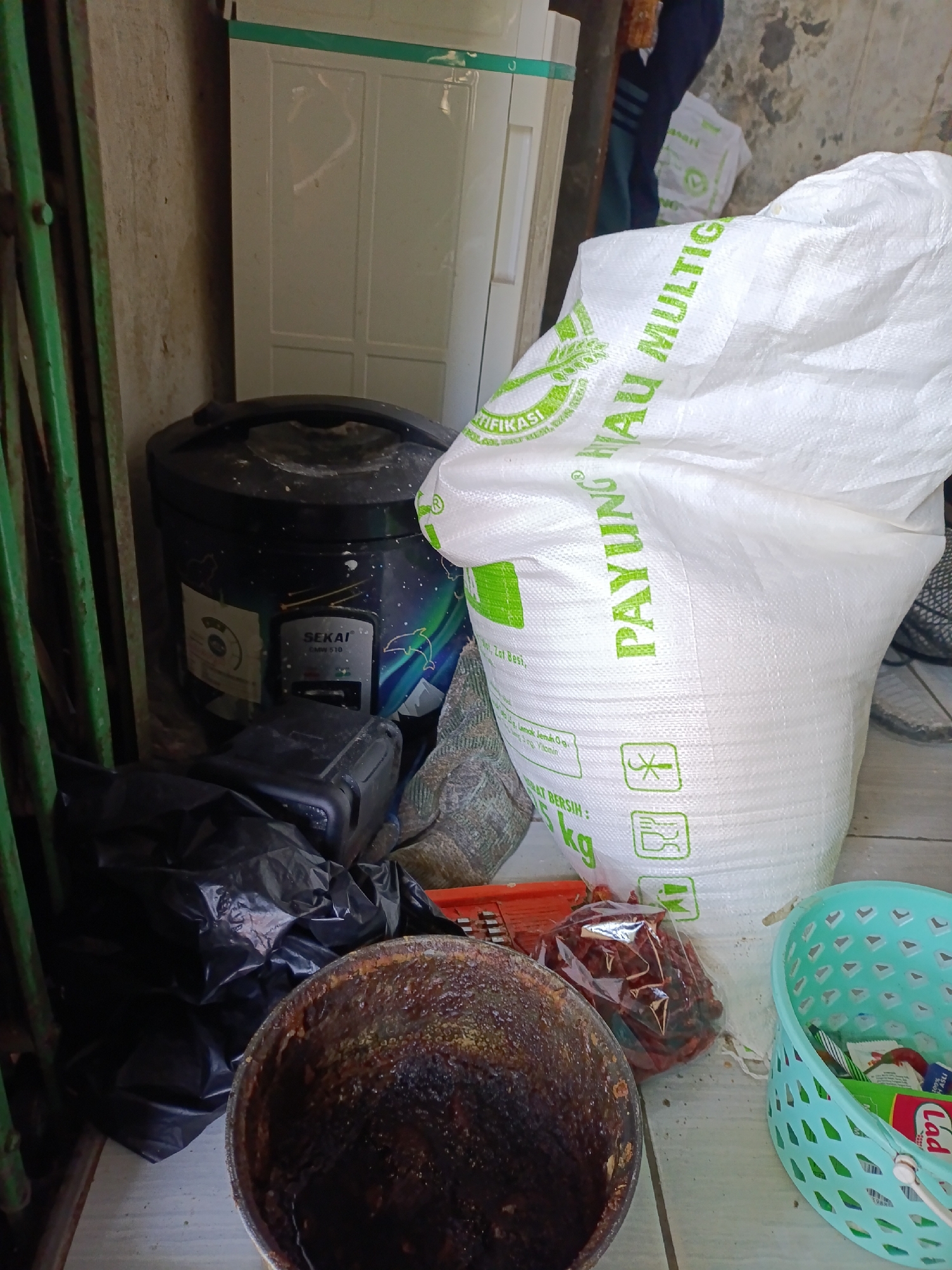 The image shows a cluttered outdoor area, possibly a storage space or a side alley in Cariu, Indonesia. In the foreground, a large, white woven bag with green lettering is prominent. The text on the bag, though partially obscured, appears to read "PAYUNG NAU MULTI" and "5 KG". To the left of the bag, a black rice cooker is visible. In front of the rice cooker are black plastic bags filled with trash. A large, dark, bowl-like object containing a dark, viscous substance sits in the lower left corner. Behind the rice cooker is a white cabinet or refrigerator. To the right of the large white bag, a blue plastic basket is filled with various items, including what appear to be small packets or wrappers. In the background, a person's legs, clad in dark pants, are visible, suggesting human presence in the vicinity. The overall environment appears unkempt, with a concrete floor and a textured, weathered wall on the left. The scene conveys a sense of everyday life and storage in an Indonesian setting.