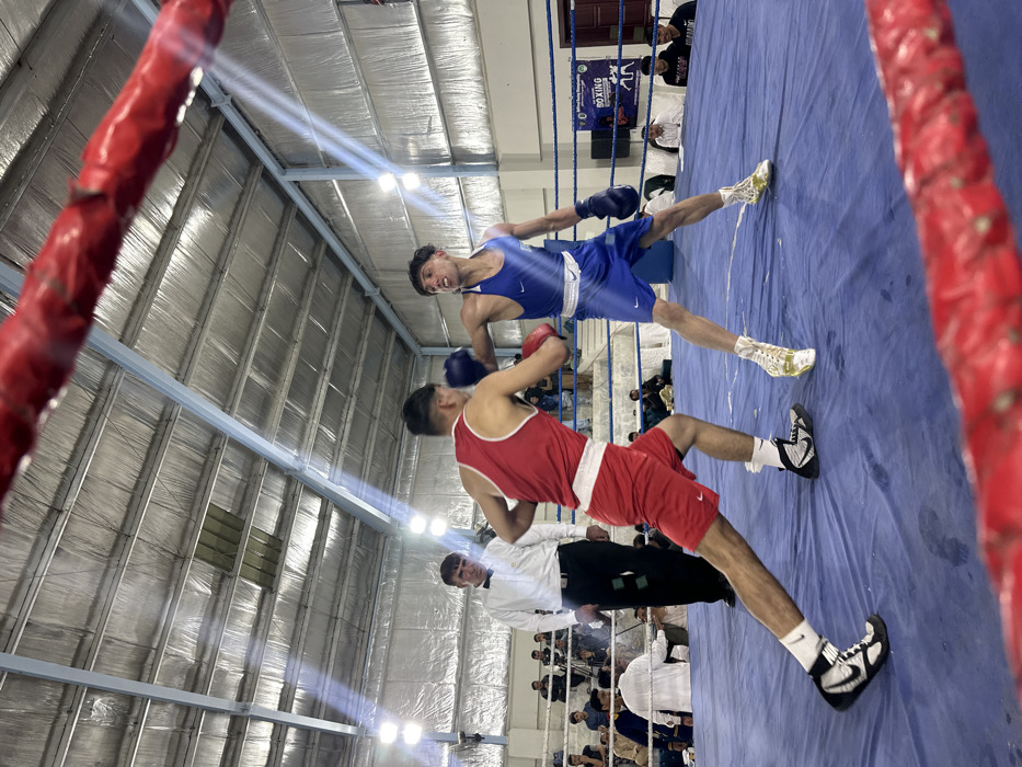 This image depicts an indoor boxing match in Quetta City Tehsil, Pakistan. Two male boxers are engaged within a blue-floored boxing ring, marked with white lines. A boxer in a red tank top, red shorts, black and white boxing shoes, and red gloves is moving forward, partially viewed from his left side. His opponent, wearing a blue tank top, blue shorts, white and gold boxing shoes, and blue gloves, faces him directly, extending his right arm in a punching motion with his blue glove. A male referee, dressed in a white shirt with a black bow tie and black trousers, stands behind the boxer in red, observing the match intently. In the background, multiple spectators and officials are seated on tiered benches around the ring. The venue features a high, corrugated metal ceiling with exposed structural beams and bright overhead artificial lighting. A blue banner promoting a "National Boxing Championship" is visible on the right side of the background. The photograph is taken from inside the boxing ring, with a padded red rope in the immediate foreground.
