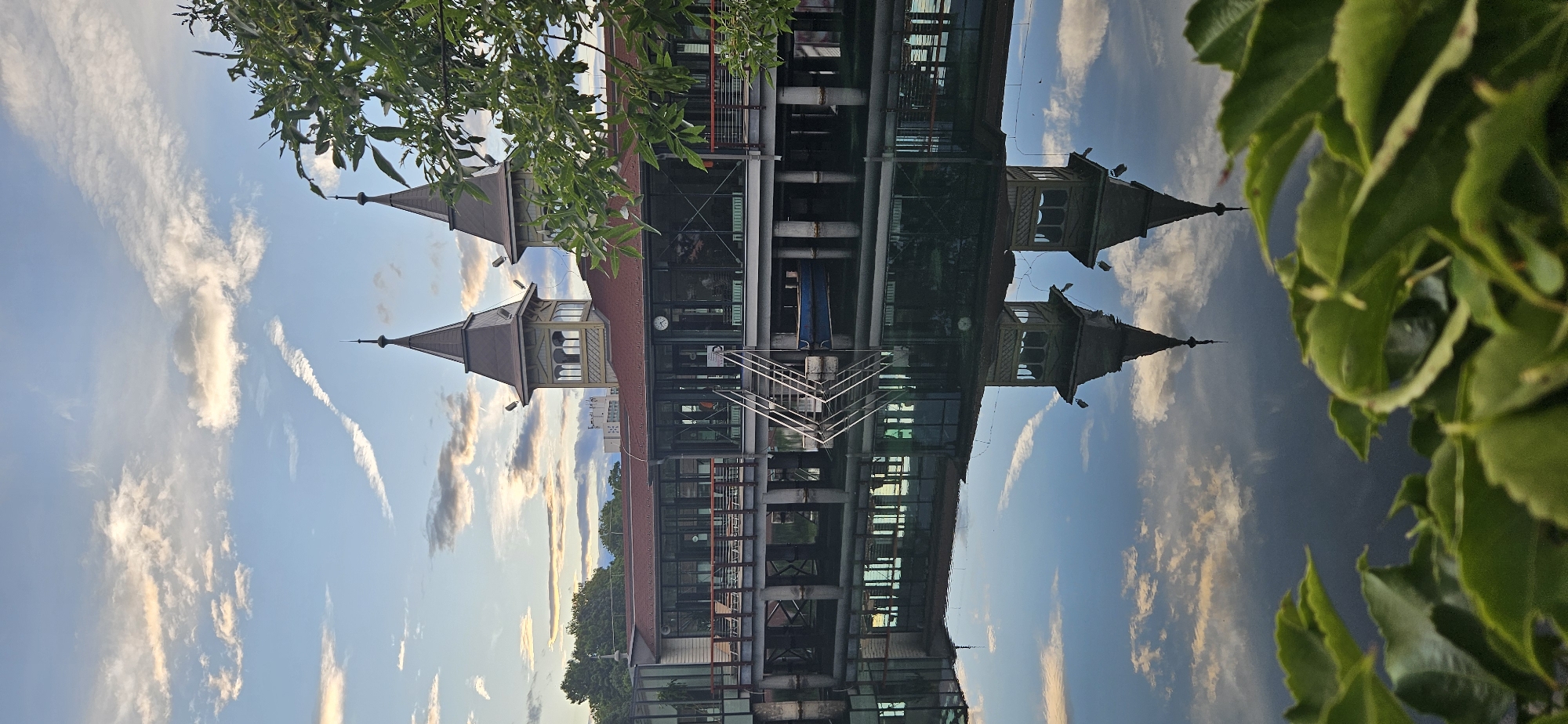 This image, taken in Hévíz, Hungary, depicts a reflection in calm water of a building with two distinct spires. The building features a red roof and a multi-story structure with large windows, metal piping, and an outdoor staircase. The spires are dark gray with pointed tops and decorative elements near the apex. The reflection is clear, showing the building and the sky above.
The sky is a bright blue with scattered white and wispy clouds, some tinged with golden light suggesting it is either early morning or late afternoon. Green leaves from a nearby plant frame the right side of the image, some in focus and others slightly blurred. The overall scene conveys a sense of tranquility, amplified by the still water and the soft lighting. No people are visible in the image. There is some text on a sign on the building's exterior, but it is too small to read clearly.