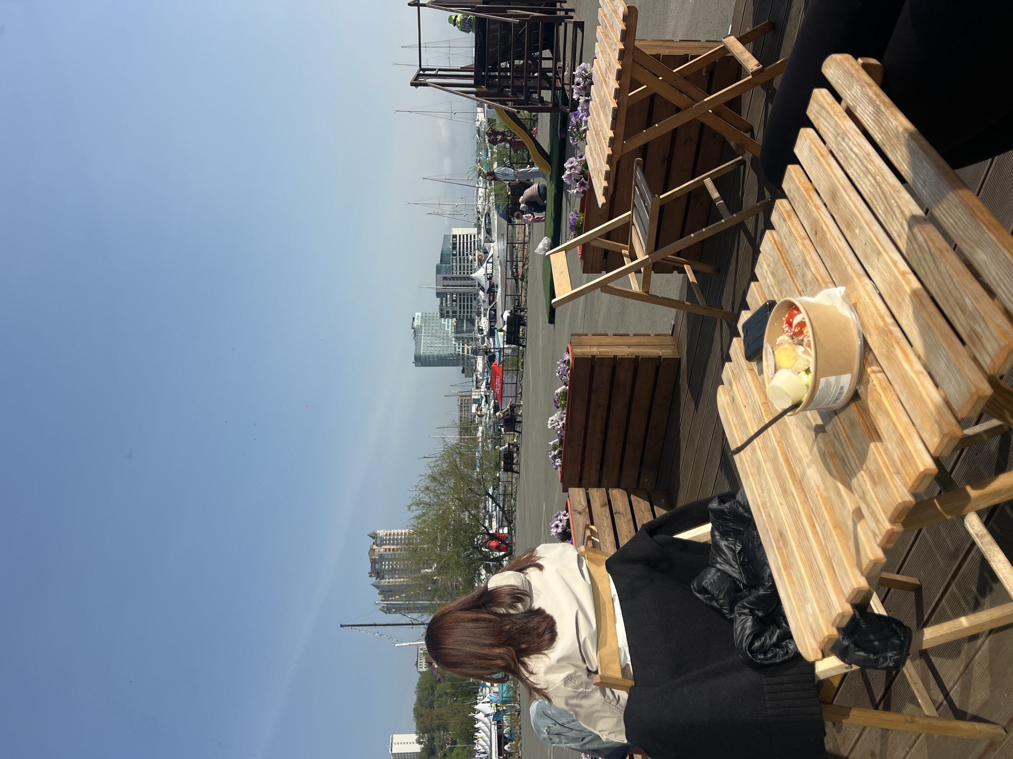 A medium-shot photo shows a scene at a marina in Vladivostok, Russia, on a clear day. In the foreground, a wooden table with a takeout container of food rests beside a person with long brown hair, wearing a light-colored jacket, seated in a wooden chair. A black garment is draped over the back of the chair. Another wooden table and chair set is nearby. In the background, the cityscape is visible with various buildings, including residential towers, and numerous boats docked in the marina. A small playground structure is also present.