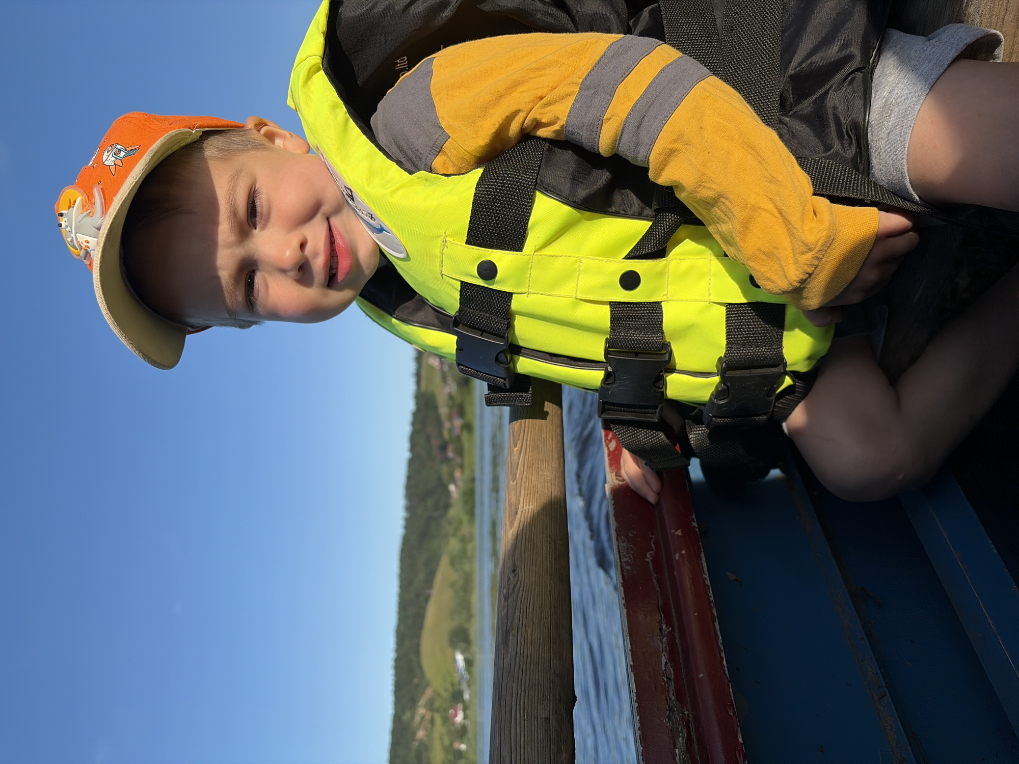 The image shows a young boy wearing an orange baseball cap with cartoon characters and a bright yellow life jacket. He is smiling and looking towards the camera. The boy is sitting on what appears to be a boat or dock, with a body of water and a tree-lined shore in the background. The sky is clear and blue, suggesting a sunny day. The overall scene evokes a sense of leisure and outdoor activity, likely on a lake or river. The location is identified as Olkhovka, Russia. No visible text is present in the image.