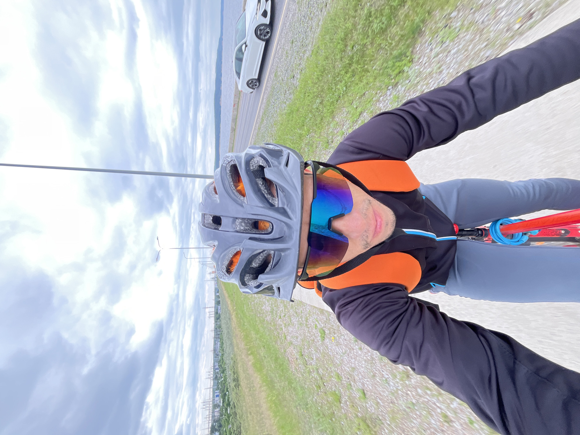 An over-the-shoulder, wide-angle selfie captures a person cycling on a paved path next to a road and a body of water, likely the Kama River near Chaikovsky, Russia. The individual is wearing a gray bicycle helmet with orange accents, mirrored cycling glasses with blue and purple lenses, a black long-sleeved cycling jersey, gray cycling shorts, and an orange backpack. The cyclist's face is visible, looking forward. To the right of the path, a white car drives on the road. In the background, a body of water stretches to the horizon, with a shoreline and scattered vegetation. The sky is overcast with heavy clouds. Streetlights are visible along the road, and distant industrial structures and bridge supports can be seen on the left side of the frame. The overall scene depicts an outdoor recreational activity in an urban or semi-urban environment with a natural water element.