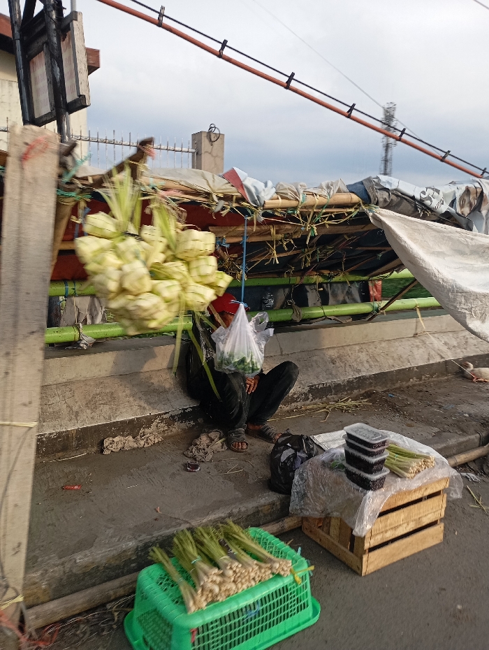 A man sits on the ground underneath a makeshift shelter constructed of bamboo and tarp, situated in a city setting in Margaasih, Indonesia (latitude: -6.973, longitude: 107.548). The man is wearing a red shirt and dark pants. He holds a plastic bag with green vegetables in it. In front of him are several containers of goods, likely for sale. A green plastic crate filled with bunches of lemongrass sits on the ground next to a wooden crate stacked with more containers. An additional plastic bag is also on the ground. The shelter is in the process of being built and is currently propped against a concrete wall.