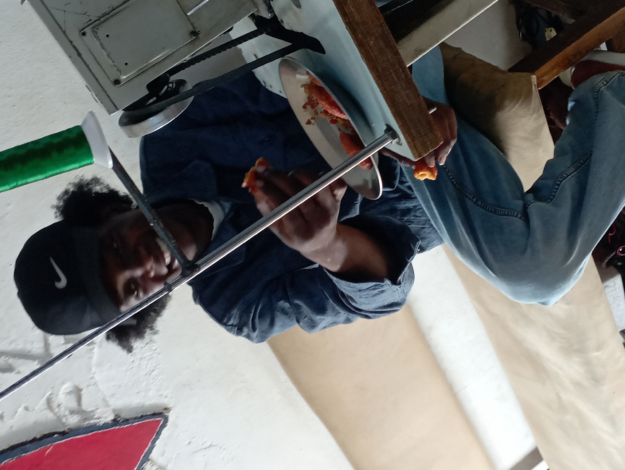 The image, taken from a low-angle perspective, captures a person, likely male, eating in what appears to be an indoor, possibly industrial or workshop, setting in Jos, Nigeria. The photograph is rotated 90 degrees clockwise; when viewed upright, the subject is looking upwards, perhaps at the camera or a ceiling structure.
The person has dark skin and curly dark hair, wearing a black baseball cap with a white Nike swoosh logo clearly visible on the front. They are dressed in a dark blue long-sleeved shirt and light blue jeans. In one hand, they hold a small piece of orange-colored food, while a metal plate resting nearby contains more of the same, partially eaten food, possibly yams, plantain, or a similar item.
Above and around the person, various elements suggest a functional or work-oriented environment. Parts of what appears to be machinery or a workbench are visible, including a gray metal box, a black belt, and a pulley or wheel. A metallic rod or pole extends across the foreground, passing between the person and the machinery. To the upper left, a green object, possibly a tool handle or part wrapped in green material, is seen. In the lower left, a geometric shape in red and black is visible on a white surface, possibly a logo or sign. The background consists of a light-colored, textured wall and large sheets of brown or tan material, possibly paper or fabric, draped over a surface.
The activity depicted is the person taking a meal or snack break. Their expression is neutral to slightly smiling, suggesting a relaxed moment. The overall scene conveys an informal, everyday moment within a practical, perhaps industrial or creative, workspace.
