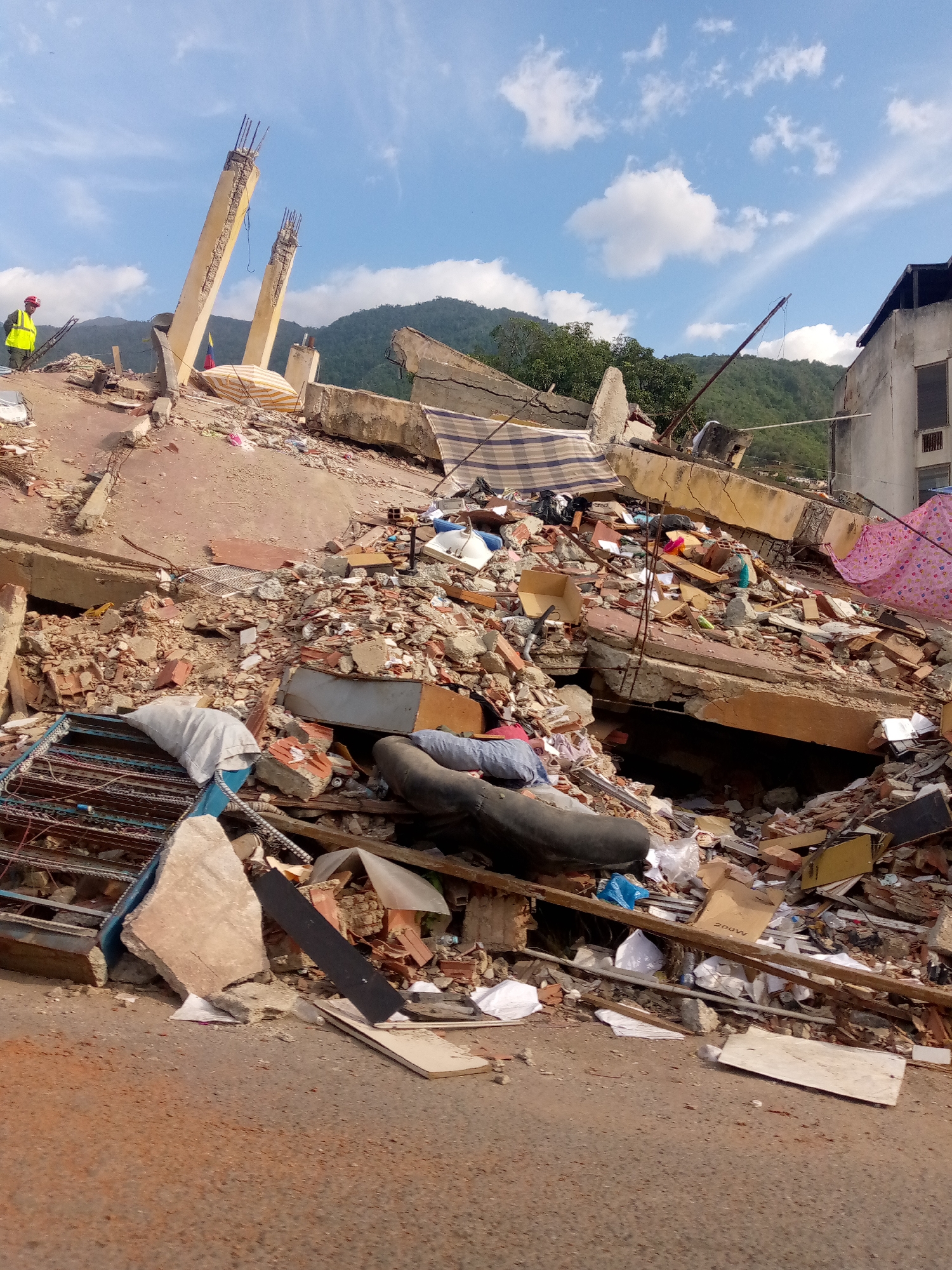 The image shows the aftermath of a disaster, likely an earthquake or landslide, in the city of Caraballeda, Venezuela. A collapsed building is the central focus, with concrete slabs, bricks, and rebar scattered across the scene. The destruction is severe, with a significant portion of the structure reduced to rubble.
In the foreground, there are various debris, including a large black tire, white bags, and a metal grate. A person wearing a yellow vest and a red helmet stands on the debris, possibly a rescuer or official assessing the situation. In the background, a blue sky with scattered clouds looms over the damaged landscape. Mountains are visible in the distance, covered in lush green vegetation. The overall scene depicts a devastating event and the challenging recovery efforts that would follow.