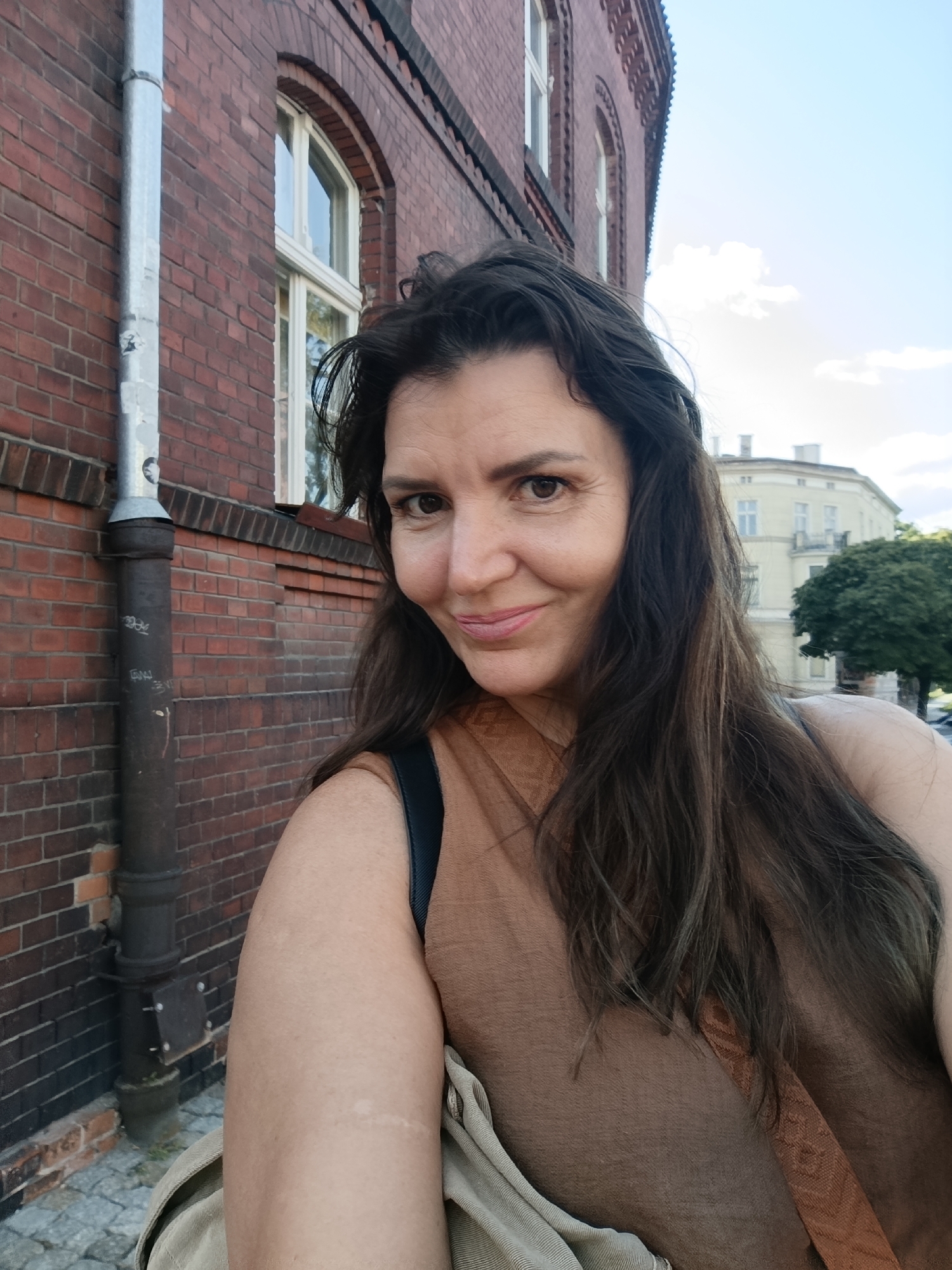 An outdoor, eye-level, medium shot captures a woman in her 40s taking a selfie in front of a red brick building. The woman has shoulder-length dark brown hair, brown eyes, and is smiling. She is wearing a tan-colored shirt and a tan backpack. To the left of the woman is a downspout attached to the brick building, which appears to be historic architecture. In the background, a light yellow, multi-story building with arched windows and a tree are visible. The sky is partly cloudy. The scene appears to be on a street in Toruń, Poland, a city known for its well-preserved medieval architecture.