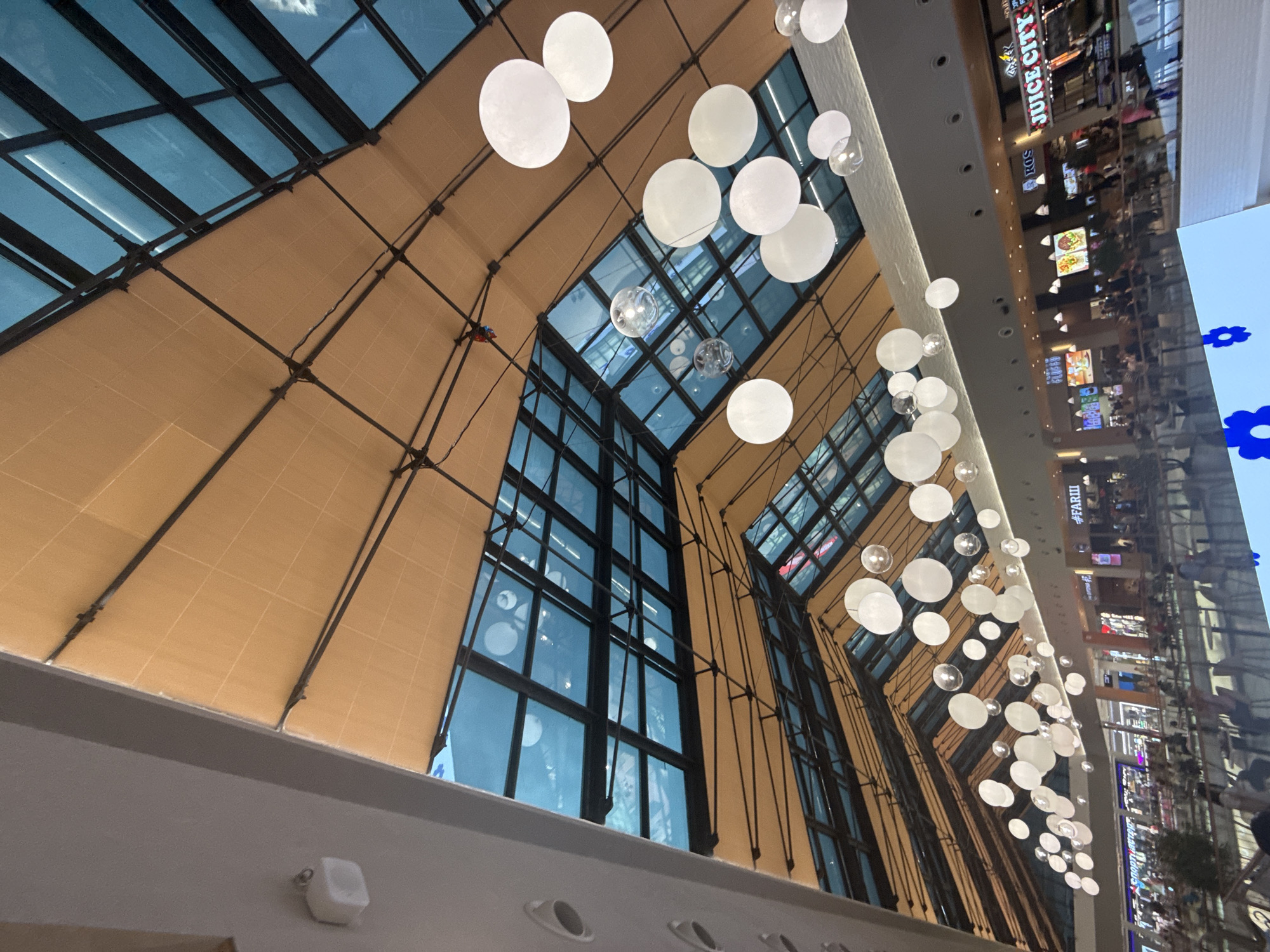 The image captures the interior of a modern shopping mall, likely the Metropolis Mall in Khimki, Russia, given the visible signage. The central focus is the high, angled ceiling, constructed with a combination of translucent tan fabric panels and large sections of blue-tinted glass, framed by dark metal beams. Numerous white, spherical light fixtures, varying in size, are suspended from the ceiling, creating a decorative element. Some smaller, clear glass spheres are also present among the larger lights. Below the ceiling, the mall's upper level is visible, with various storefronts and eateries lining the walkway. Signs such as "JUICE CITY" and "#FARIII" are discernible. Numerous people are present on the upper level, appearing to be shoppers or diners. They are seen walking, sitting at tables, and interacting with the environment. The overall impression is of a bustling, contemporary commercial space with a distinctive architectural design.