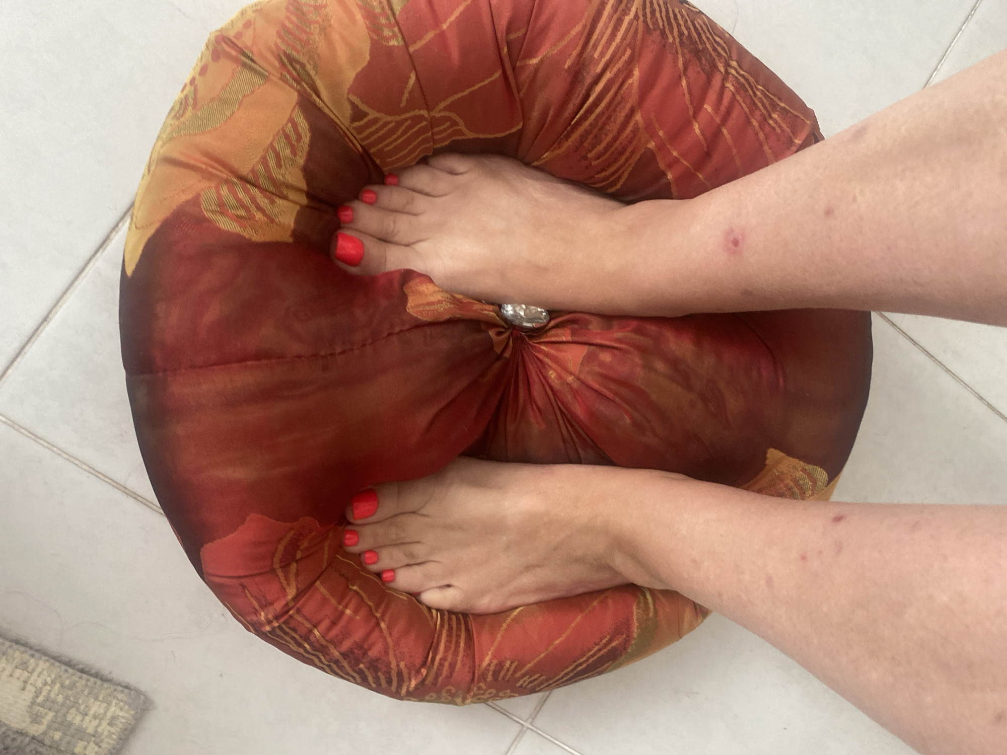 The image shows a top-down view of a person's feet and lower legs resting on a round, red and orange patterned cushion. The toenails are painted a bright neon red. The person's skin appears to have some small red marks or blemishes. The cushion is placed on a tiled floor. A grey and white patterned rug is visible in the upper left corner of the image. The scene is indoors. The location of Novi Petrivtsi, Ukraine, is not directly depicted or implied by the visual elements in the image.