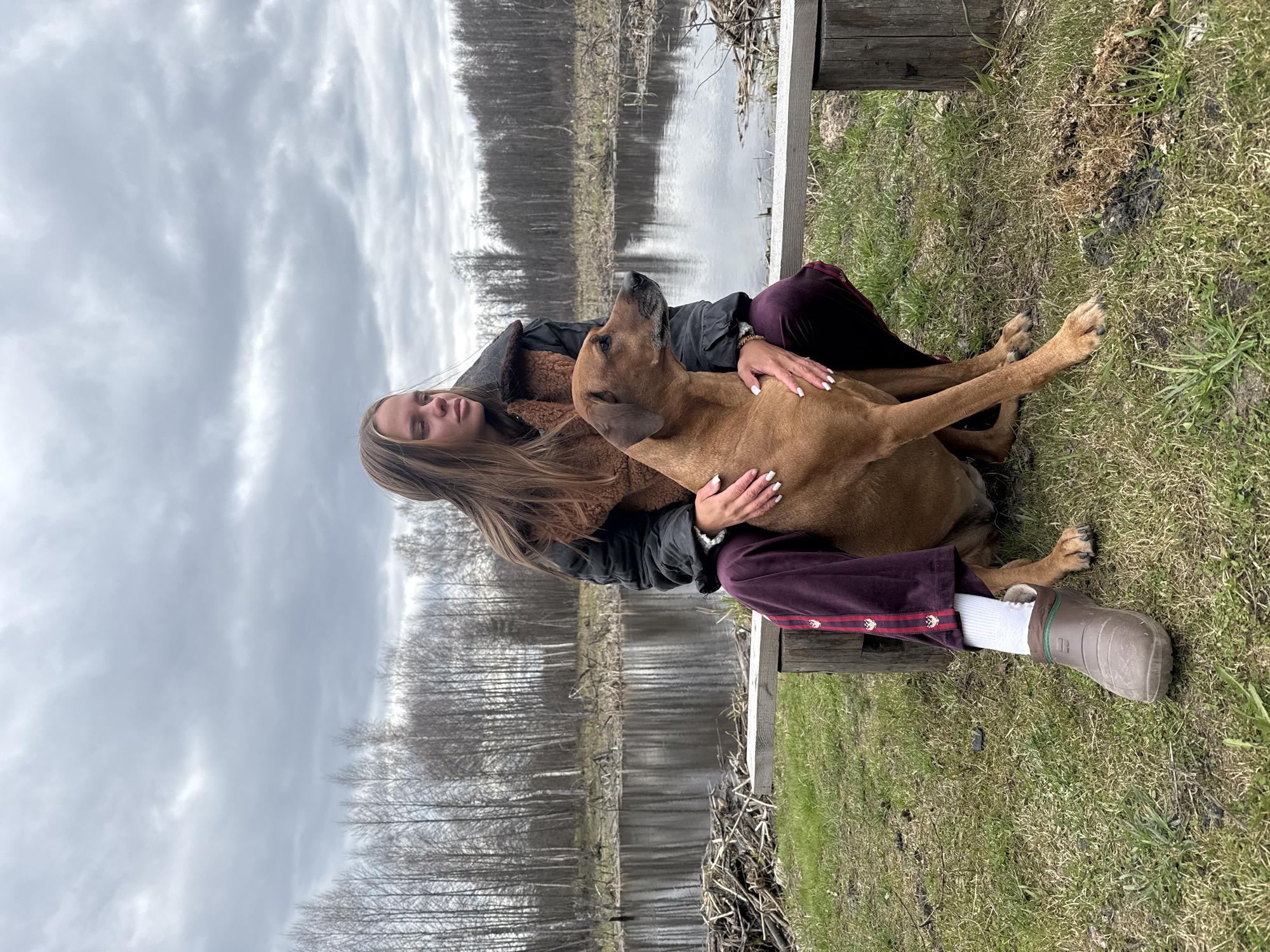 A woman with long brown hair sits on a wooden bench near a body of water in Grebnevo, Russia. She wears a black jacket over a brown sherpa-lined garment, purple velvet pants with a decorative stripe, and a single brown boot, paired with a white sock. The woman is embracing a brown dog, which is looking upwards. The background features a lake, bare trees, and an overcast sky. The ground around the bench is covered in grass and dry vegetation.