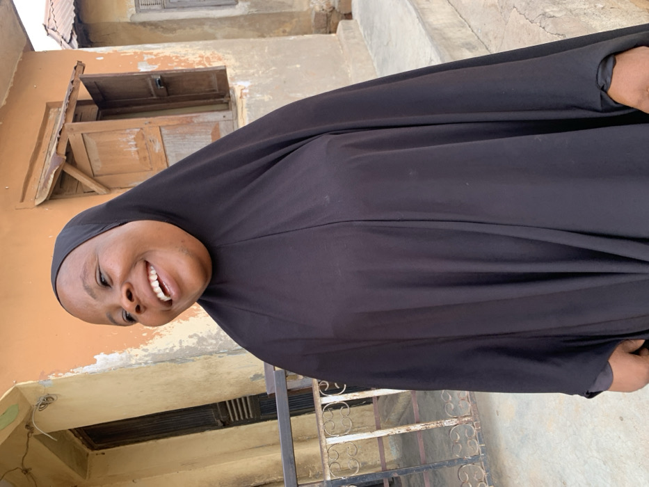 A woman wearing a black hijab smiles broadly. She is standing outdoors in front of a building with a weathered, light brown exterior. There's a window with closed shutters visible above her. The setting appears to be a residential area. A metal fence-like structure is at the bottom left corner. The image is oriented vertically.