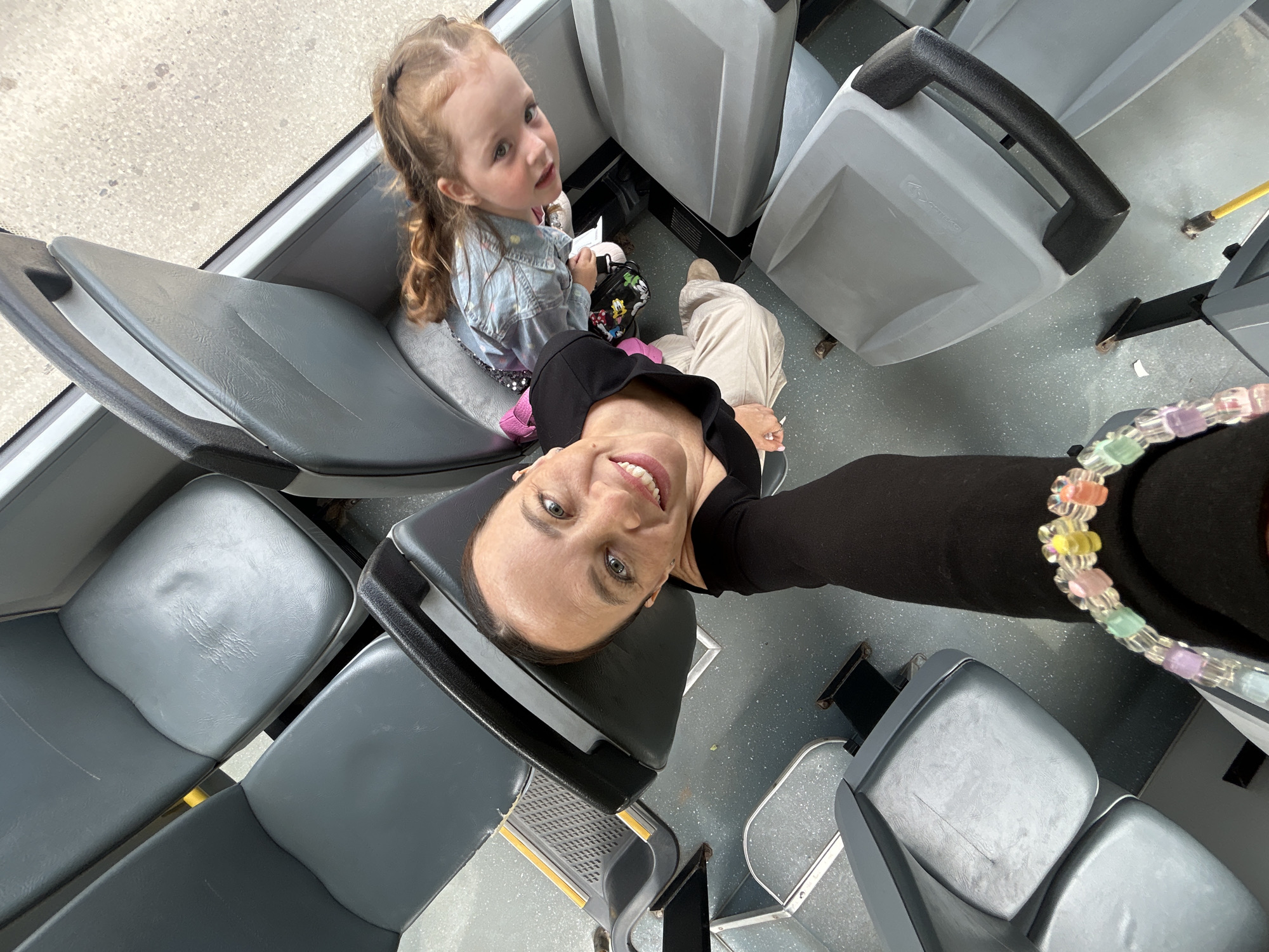 An outdoor, high-angle, full shot shows two females seated on a bus, with the camera pointing downwards. On the right, a young girl with blonde hair styled in braids, wearing a denim jacket and a pink and white polka dot skirt, sits on a gray seat. To the left, a woman with brown hair pulled back and wearing a black long-sleeved shirt and tan pants smiles at the camera. She has a colorful beaded bracelet on her left wrist. The bus interior features rows of gray plastic seats. The flooring is a speckled gray. The scene appears to be in Chaikovsky, Russia.