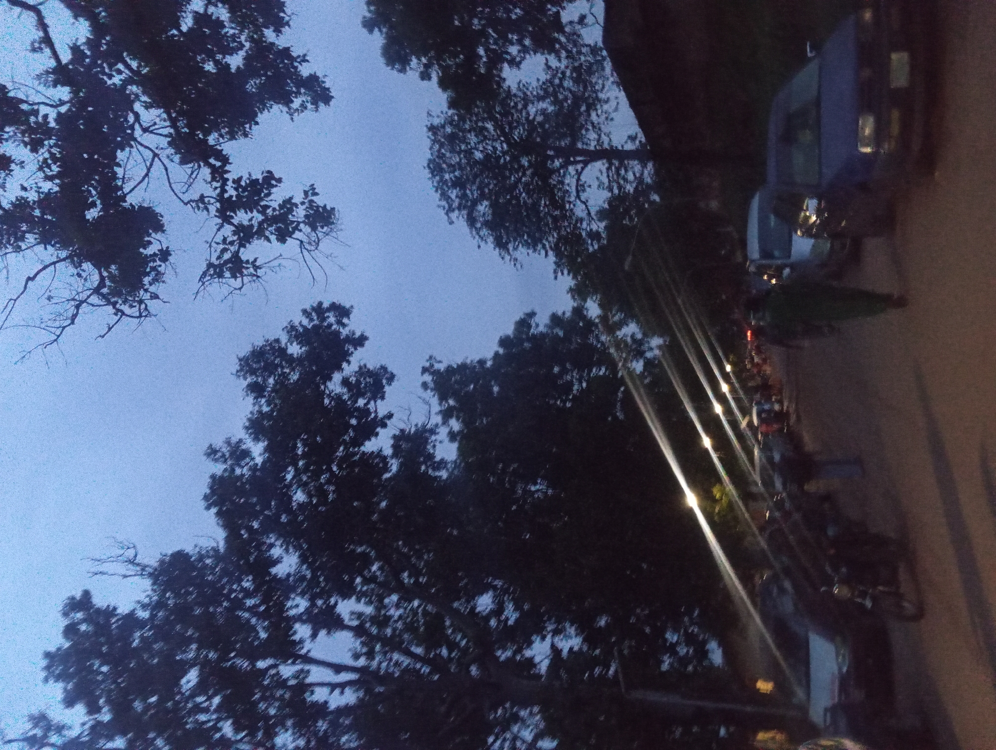 The image depicts a street scene at dusk, characterized by dim lighting and silhouettes of trees against a twilight sky. Several vehicles, including cars and a motorcycle, are parked or driving along a dirt road. People are visible, some walking and others standing near the vehicles. The environment appears to be a semi-urban or suburban area, with streetlights casting long streaks of light across the road. The overall atmosphere is that of an evening gathering or activity taking place outdoors.