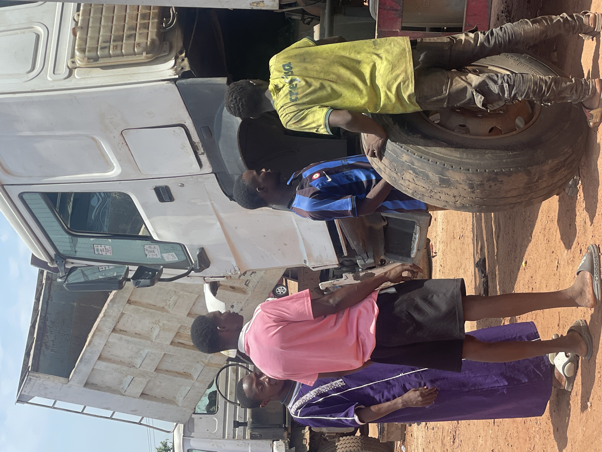 The image depicts a group of four African men working on a large, white truck in Kwali, Nigeria. Two men are focused on the truck's tire, one wearing a yellow shirt with "CERESER" written on the back and the other in a blue and black striped shirt. The other two men are standing in the front of the truck, one in a pink shirt and the other in a purple robe. The environment appears to be an outdoor workspace with a dirt ground.