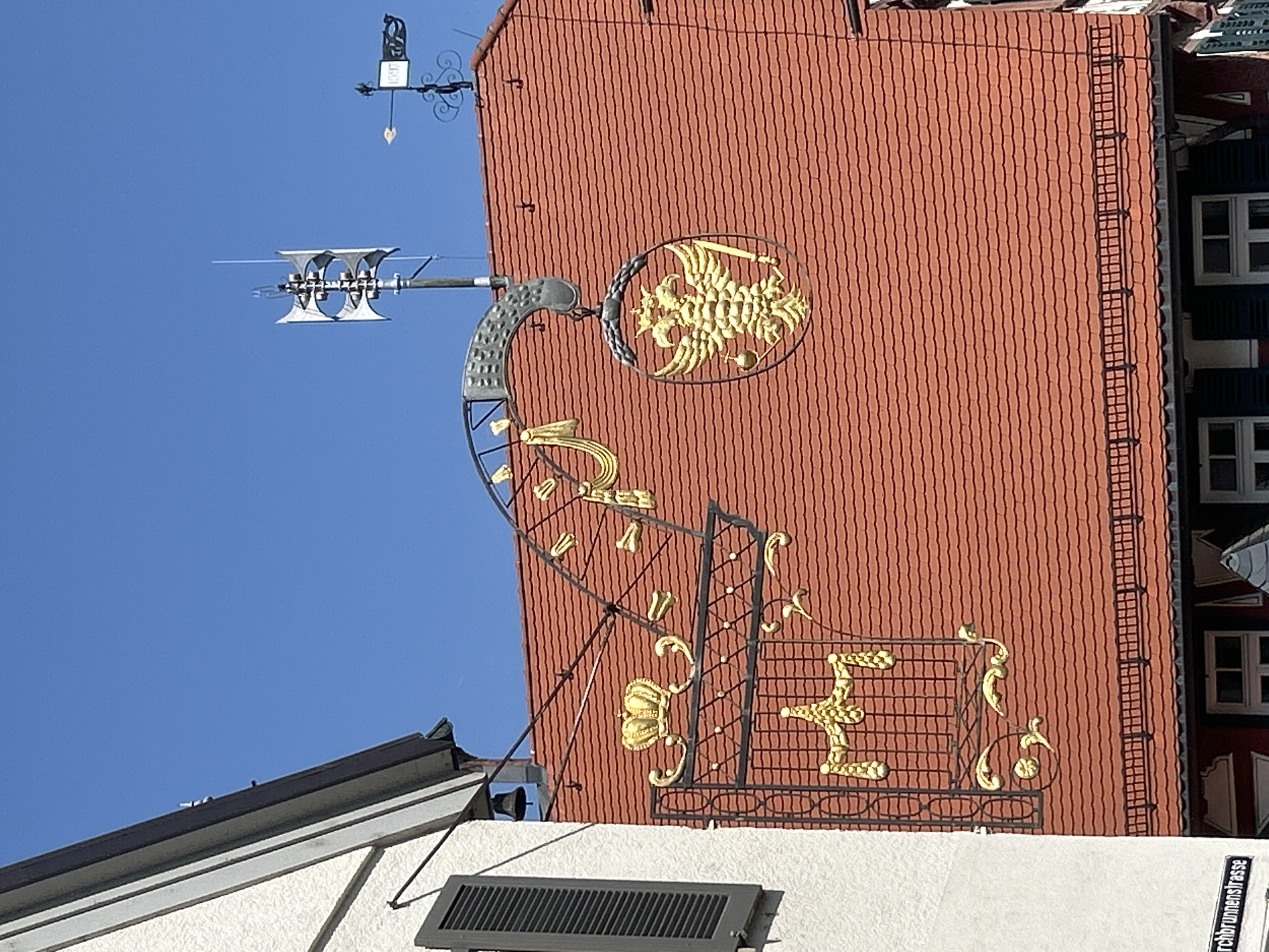 This image captures a portion of a building's facade in Bad Friedrichshall, Germany, under a clear blue sky. The main focus is a decorative wrought-iron sign hanging from the side of the building, featuring golden embellishments. The sign has an ornate, curved arm extending from the building, from which a circular plaque hangs. This plaque depicts a detailed golden double-headed eagle with outstretched wings, holding a sword in one talon and an orb in the other. To the left of the eagle plaque, a golden crown sits atop a decorative grid structure. The building itself has a red-tiled roof. In the background, a weather vane and a siren are visible against the sky. Part of an adjacent white building with a dark shutter is also visible on the left. A street sign partially visible at the bottom left reads " Kirchbrunnenstrasse".