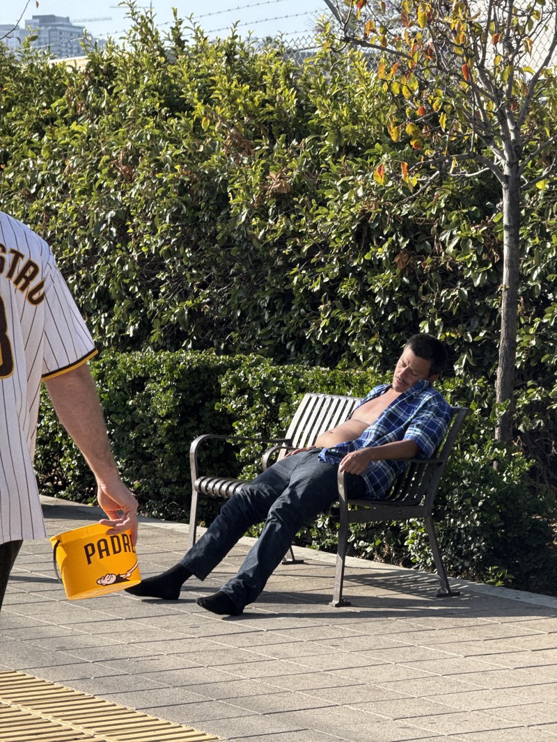 A man is sleeping on a park bench on a paved area near a large hedge and a thin tree. He is wearing black socks and is barefoot, dark denim jeans, and a blue plaid shirt. The shirt is open, and his bare chest is visible. An individual wearing a baseball jersey that is predominantly white with vertical pinstripes and a tan shoulder is holding a yellow bucket with the word "PADRES" in black capital letters. The setting appears to be outdoors in San Diego, United States.
