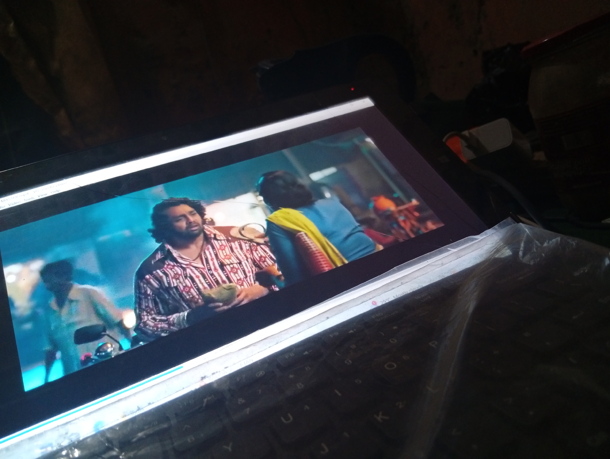 The image shows a laptop screen displaying a scene from a movie or TV show. On the screen, two men are visible. The man in the foreground is wearing a striped shirt and holding a green object, possibly a fruit or vegetable. He has dark, curly hair and a beard. The second man is in the background, on a motorcycle, wearing a light-colored shirt. The screen is illuminated by a bright blue light. The laptop itself is placed on a keyboard, which is partially covered by a clear plastic sheet. The background of the image is dark and out of focus, suggesting an indoor environment. There is no identifiable information about the location within the city of Unknown.