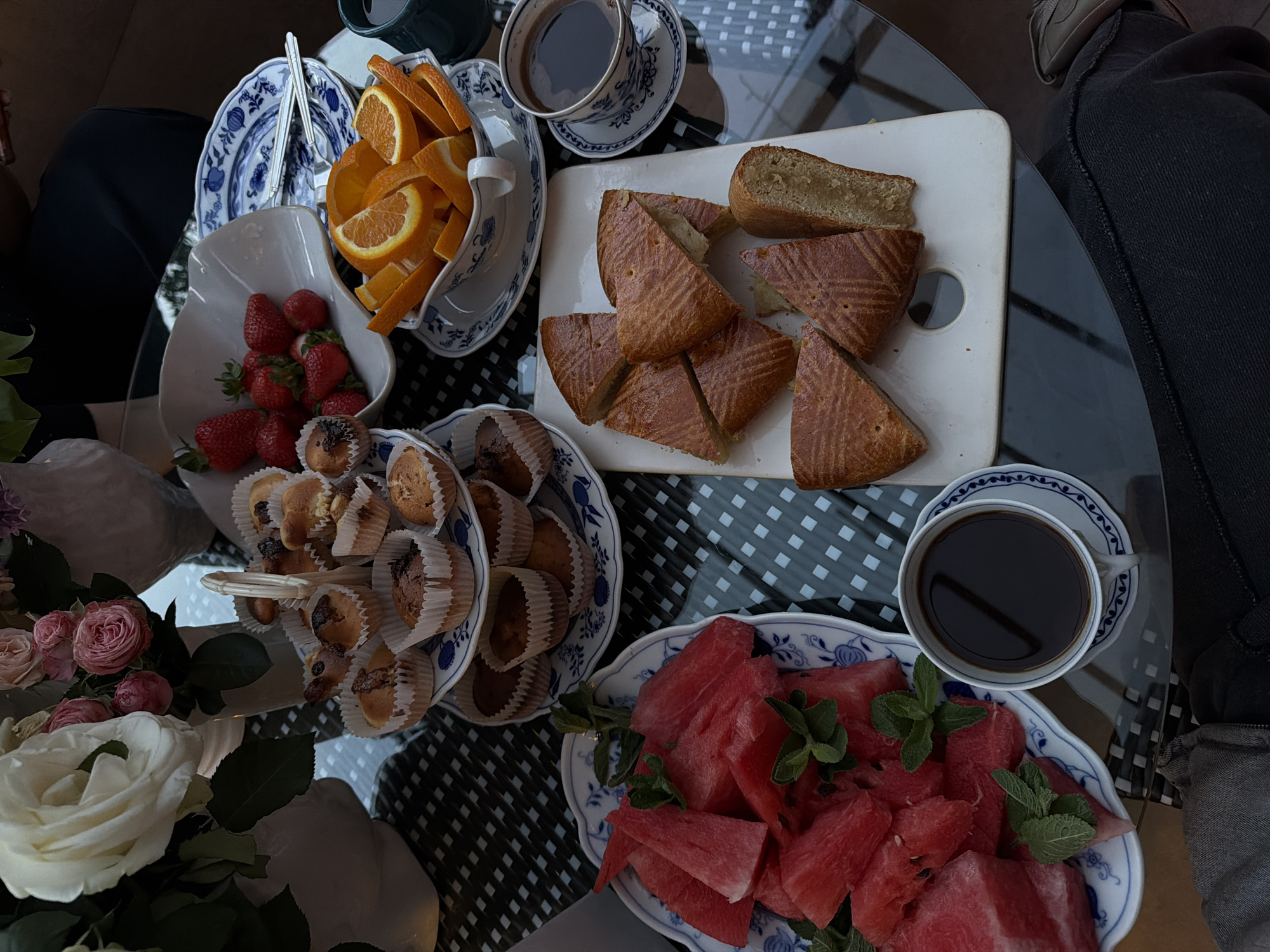 A top-down shot shows a glass-topped table laden with food and drinks. There are oranges in a white-and-blue bowl, next to a white bowl of strawberries. A tiered white-and-blue serving dish holds cupcakes. A white cutting board displays cut triangles of pastry and a slice of bread. There are two cups of coffee. Watermelon pieces with mint leaves fill a white-and-blue dish. A bouquet of white and pink roses sits near the left edge. The table is covered with a checkered black-and-white tablecloth. A person wearing dark pants and a light-colored shoe sits to the right. The location is most likely not in Yerevan, Armenia due to the lack of contextual information, which is outside the bounds of the objects in the image.