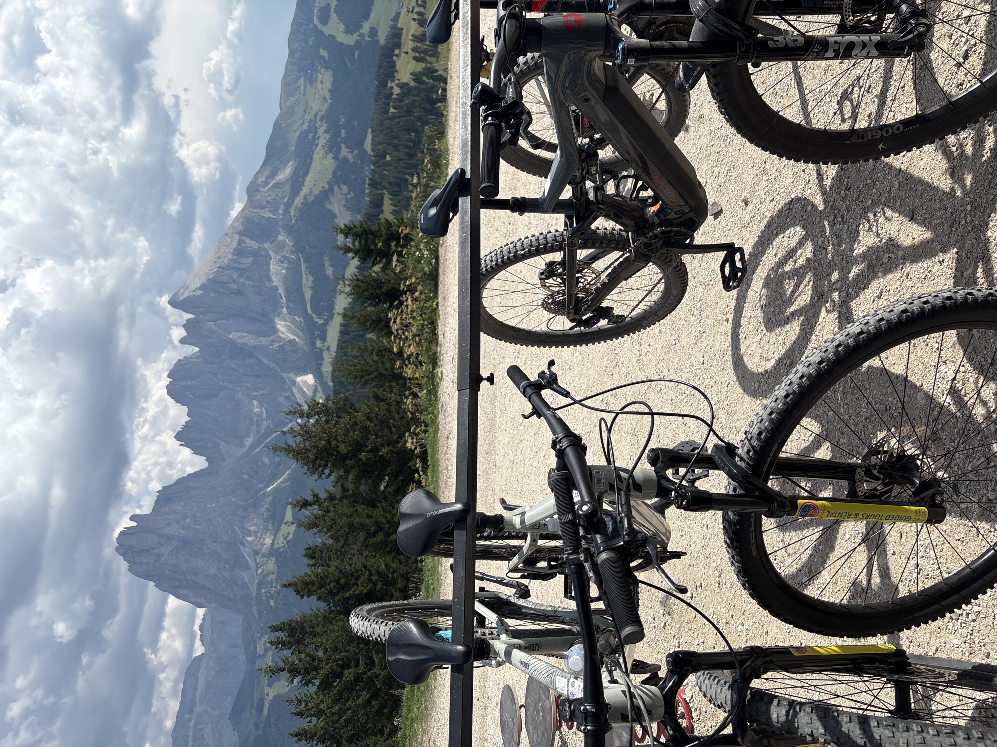 The image shows several mountain bikes parked on a gravel surface, with a mountain range and cloudy sky in the background. The bikes are positioned in a rack, with some hanging and others resting on the ground. The foreground is dominated by the front wheels and handlebars of the bikes. In the background, a dramatic mountain landscape with sharp peaks is visible, partially obscured by clouds. Pine trees dot the slopes of the mountains. The overall scene suggests an outdoor recreational area, possibly a bike rental station or a parking area for hikers and cyclists in a mountainous region like the Dolomites, which is near Kastelruth - Castelrotto, Italy.
