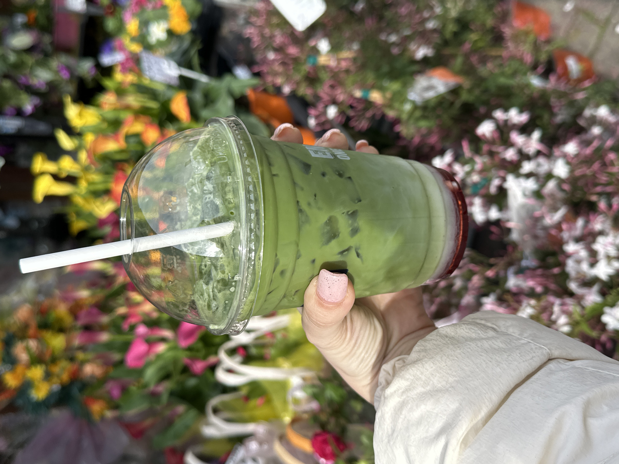 A hand holds a large, clear plastic cup filled with a green liquid, likely a matcha latte with ice, against a vibrant floral background. The cup has a dome-shaped lid and a white straw. The hand has painted fingernails with a light pink color. The floral background consists of various colors including yellow, pink, orange, purple, and white, suggesting the presence of flowers in a shop or market. The arm is wearing a light-colored jacket with white fabric. The image appears to be taken outdoors, possibly at a flower stall in London, UK, though without additional context, this is based on the common association of flower markets with the city.