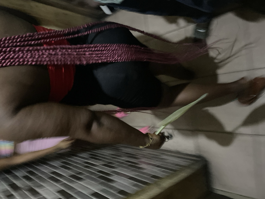 In Alaiyabiagba, Nigeria, a partial interior scene depicts two individuals within what appears to be a tiled corridor or room. The primary subject, seen from behind, is a person with dark skin, wearing a red strapless top, black form-fitting shorts, and long, burgundy box braids extending down their back. A gold-colored bracelet is visible on their right wrist. This individual holds a single, elongated light green leaf in their right hand and wears brown low-heeled footwear on their right foot, appearing to be in motion. To the immediate left of this subject, a wall is tiled with light-colored, rectangular tiles arranged in a diagonal pattern. Further in the background, a second, highly blurred individual is partially visible, wearing a light pink garment and a multi-colored (rainbow-striped) top. The floor is light-colored and tiled. The scene is dimly lit, casting shadows.