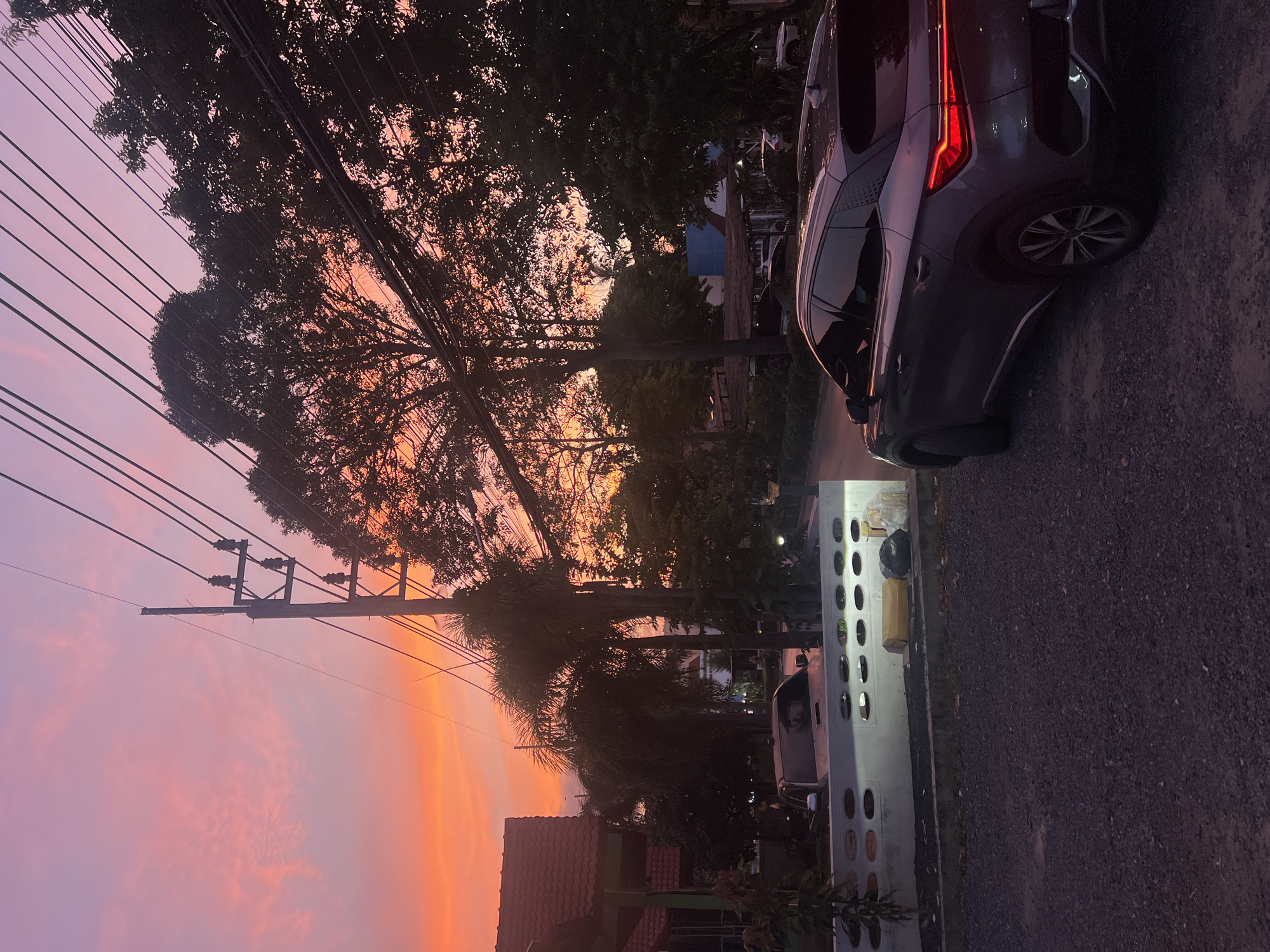 The image depicts a street scene in the city of Ko Samui District, Thailand, during a sunset. The sky is a vibrant gradient of orange and pink hues, with scattered clouds. Power lines and a utility pole are prominent in the frame, stretching across the sky and silhouetted against the colorful sunset. Several trees, including palm trees, are also visible, their foliage dark against the bright sky.
In the foreground, a dark-colored SUV is parked on a gravel surface. Its taillights are illuminated. To the left of the SUV, a white concrete wall with oval-shaped openings is partially visible. A yellow box and a black trash bag are placed near this wall. Further back, a house with a red tiled roof can be seen. The overall atmosphere is quiet and serene, characteristic of a residential area at dusk. The image is taken from a low angle, emphasizing the dramatic sky.