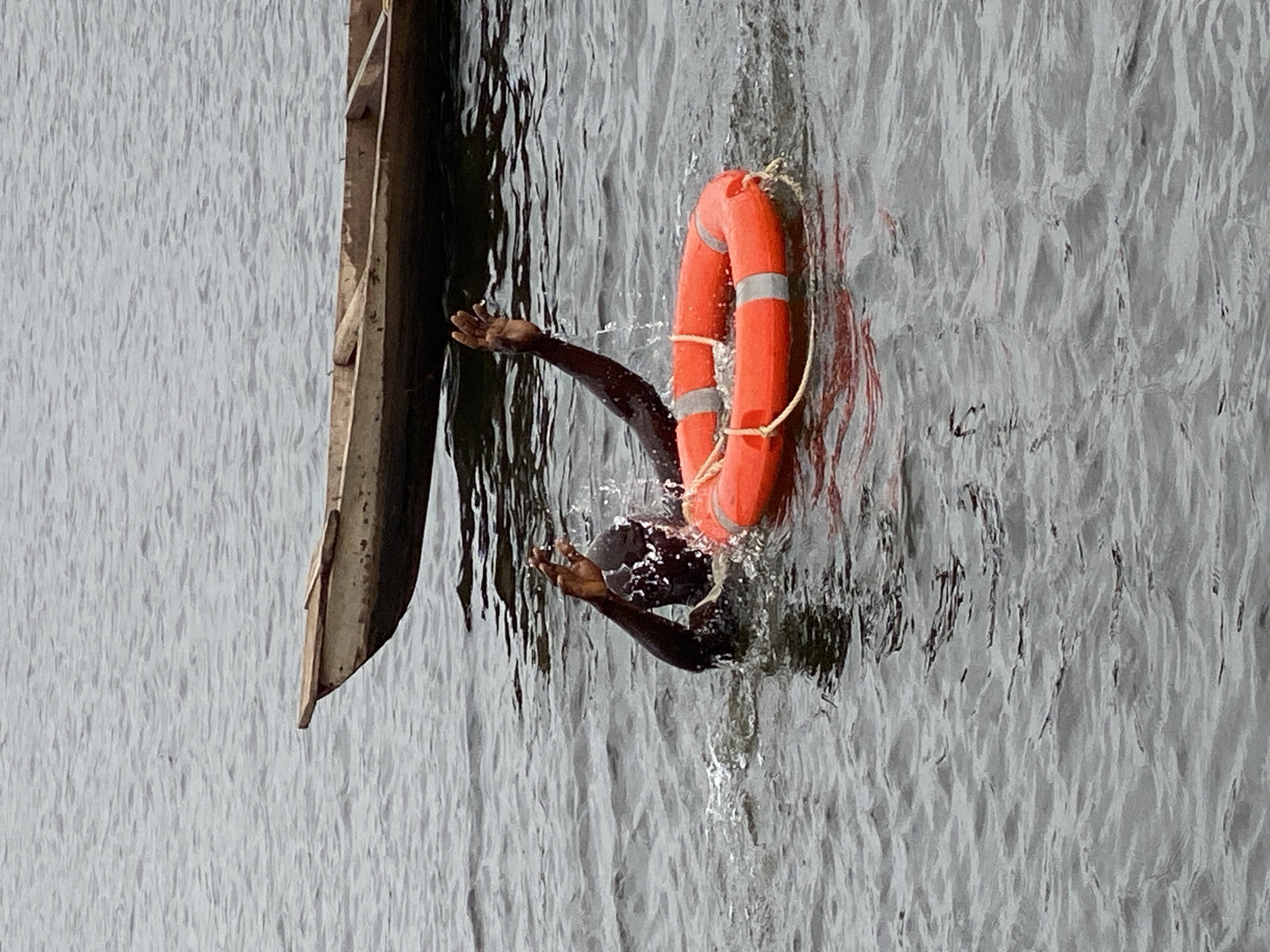 The image depicts a person struggling in dark, choppy water near a small wooden boat. The person, with dark skin, is partially submerged and has their arms raised, seemingly reaching out for help. A bright orange life preserver with reflective silver bands is floating nearby in the water. The boat, a narrow wooden vessel, is positioned in the background, partially out of focus, with its front end angled slightly upwards. The water appears to be a river or a lake, with ripples and small waves on its surface. The lighting suggests an overcast day. The scene appears to be taking place on the riverbank in Kawo, Nigeria.