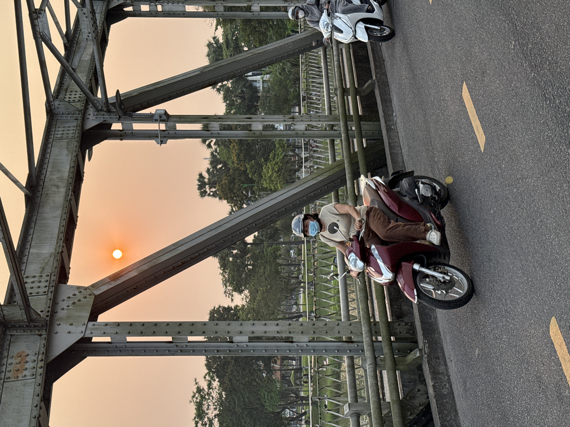 The image captures a scene on a bridge in Phú Xuân Ward, Vietnam, during sunset. An orange sun shines through the bridge’s steel truss structure. Two people are riding motorbikes. One person, wearing a helmet and a surgical mask, is riding a red scooter. Further ahead is another scooter, with two riders, partially visible. Yellow lines mark the road on the bridge. Trees and buildings are visible in the background.