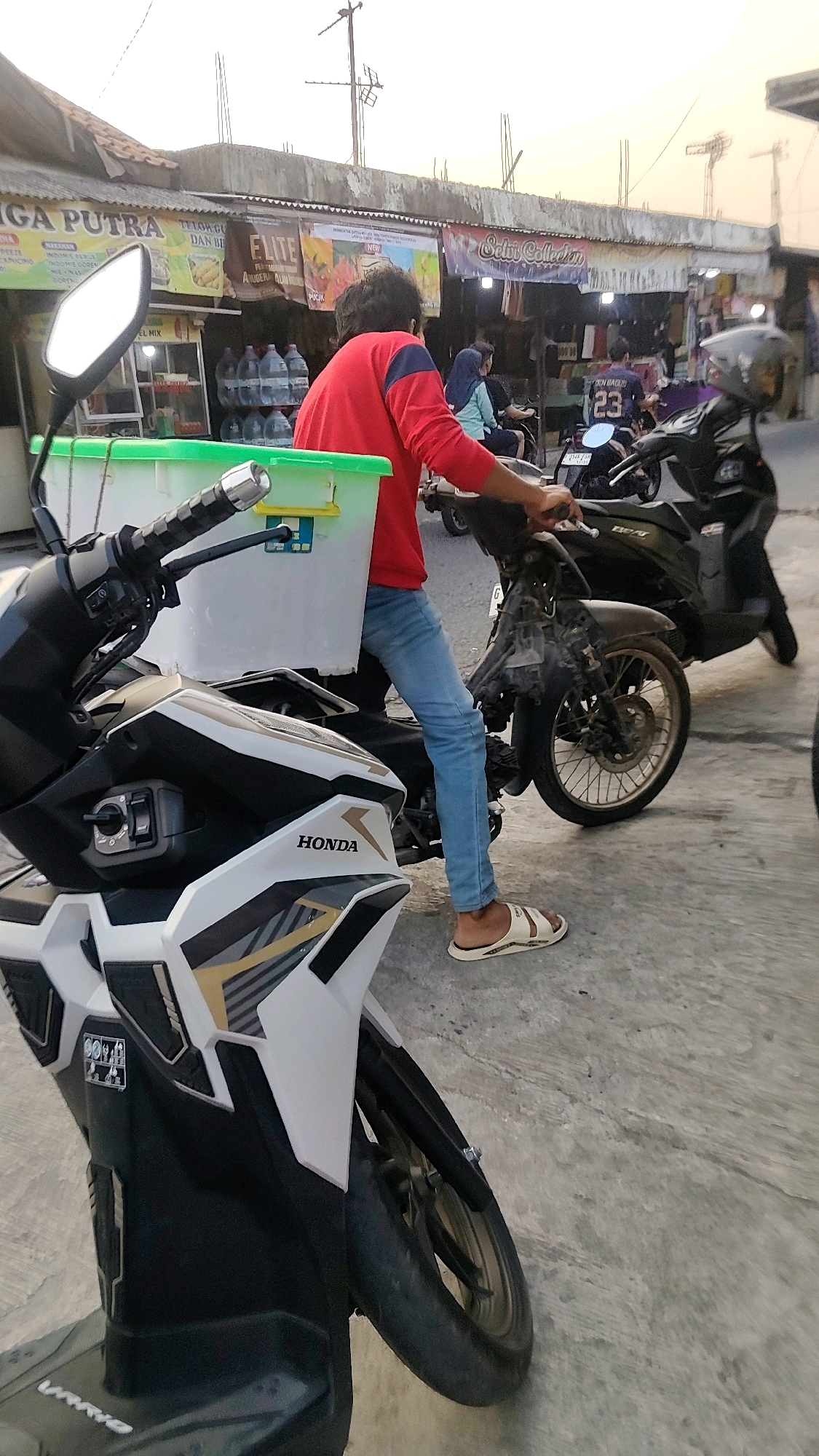 In Mekarmukti, Indonesia, a man wearing a red long-sleeved shirt and blue jeans, along with white sandals, is seated on a dark-colored motorcycle. He is holding the handlebars and appears to be either starting the engine or waiting to depart. A large white plastic container with a green lid is attached to the front of his motorcycle, secured by what seems to be a rack. The motorcycle is parked on a concrete surface, which shows signs of wear and tear. In the background, several other motorcycles are visible, some with riders. A row of small shops or stalls lines the street, with awnings and signage indicating various businesses. One sign displays "FLITE" in large letters, and another reads "Sikio Collection." Bottles of water and other goods are displayed outside the shops. The sky above is overcast with a light, hazy glow, suggesting either early morning or late afternoon. The overall scene depicts a typical street view in a commercial area of a town in Indonesia.