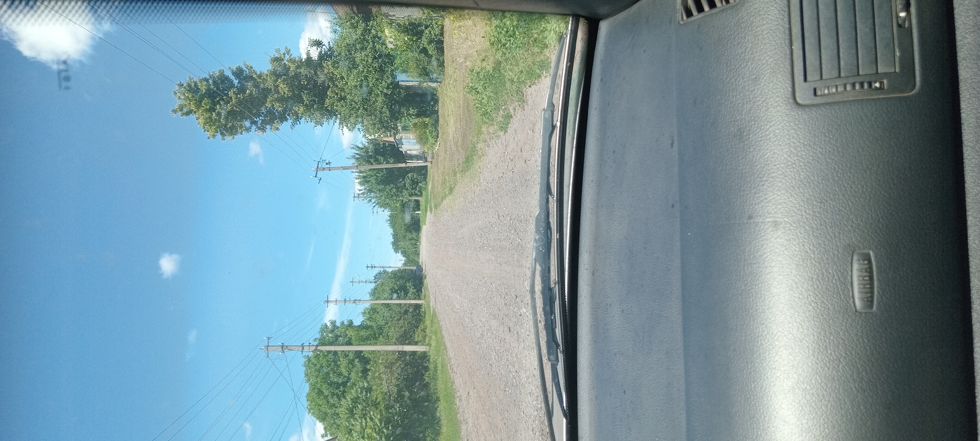 The image shows a view from inside a vehicle, looking out through the windshield at a dirt road in a rural area. The road is flanked by trees and power lines. The sky is blue with a few scattered clouds. The dashboard of the car is visible in the foreground, with the word "AIRBAG" embossed on it. The location is described as Koviahy, Ukraine, suggesting a context of everyday life in a Ukrainian village or town. The scene is peaceful and ordinary, depicting a common sight of travel through a rural landscape.