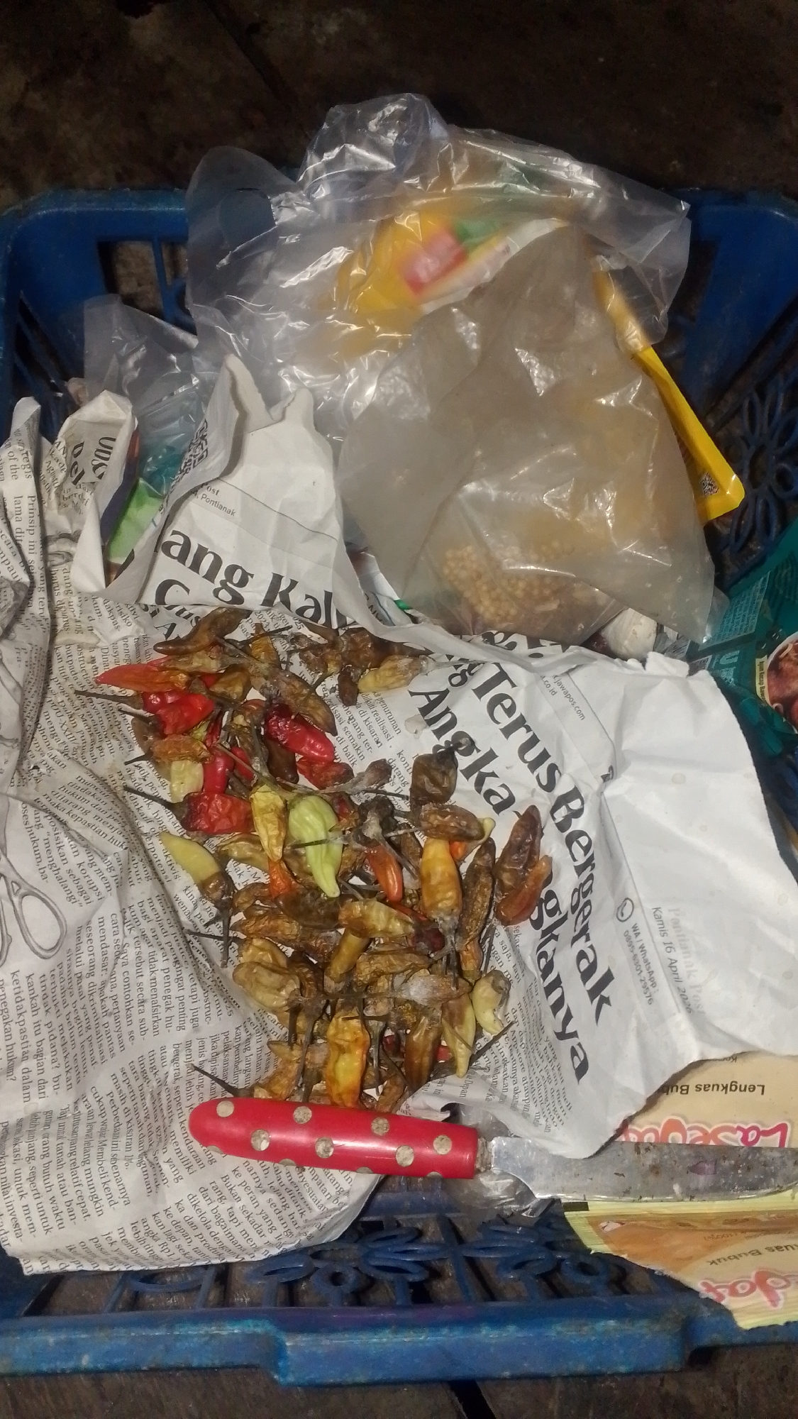 A close-up, high-angle shot captures a blue plastic basket filled with various items. In the center of the basket, a pile of dried chili peppers, ranging in color from red to green to brown, sits on top of crumpled newspapers. To the right of the chilies, a red-handled knife with a silver blade is visible. Behind the chilies, a clear plastic bag contains a yellowish substance, possibly grains, and other assorted items are loosely placed within the basket, including another clear plastic bag and parts of what appear to be food packaging. The background is dark and indistinct, suggesting an indoor or dimly lit outdoor environment. The scene appears to be located in Olak-Olak Kubu, Indonesia, based on the text visible on the newspaper, which includes Indonesian language and a possible dateline.
