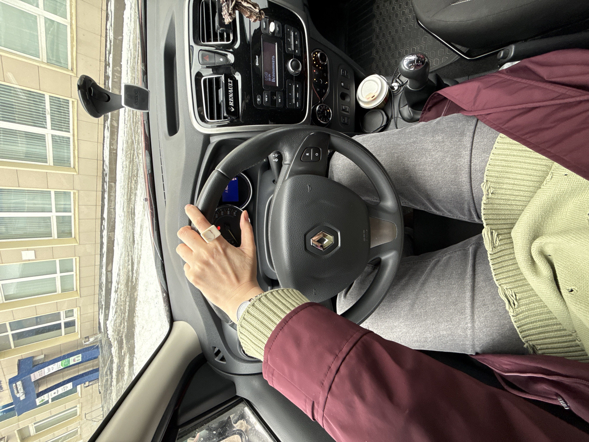 The image shows the interior of a Renault car, specifically the dashboard, steering wheel, and part of the driver. The steering wheel has the Renault logo. The driver's left hand is visible on the steering wheel, wearing a ring and a watch. There is a cup of coffee and a gear shift visible on the right side of the image. The driver is wearing gray pants, a green sweater, and a maroon jacket. Through the windshield, a building is visible, reflecting a blue sign indicating that the location is Izhevsk, Russia.