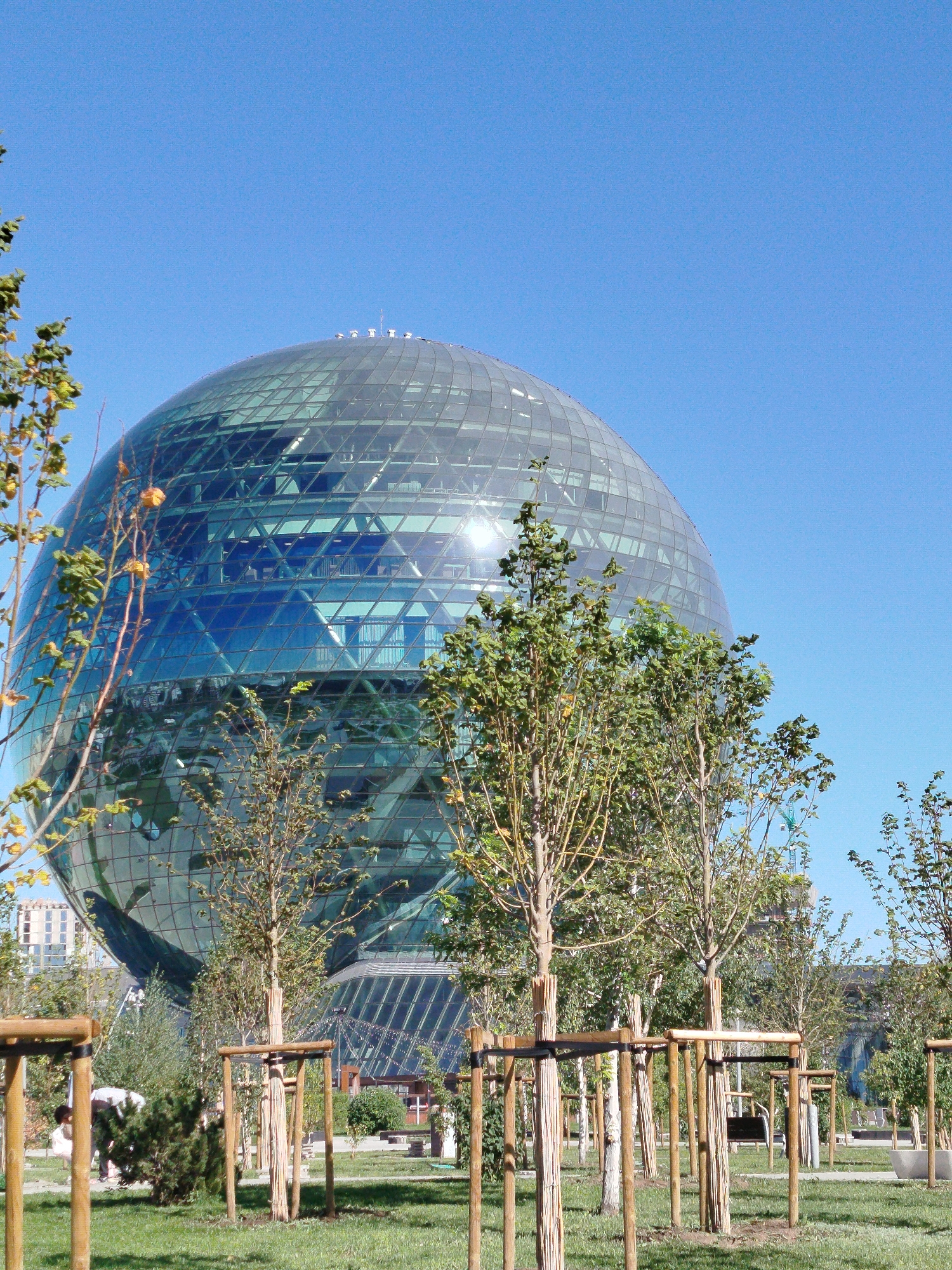 The image shows the Nur Alem pavilion, a spherical building located in Astana, Kazakhstan. The pavilion is covered in glass and has a geometric pattern. It is surrounded by a park with young trees and grassy areas. The sky is clear blue. In the foreground, there are several young trees supported by wooden stakes. In the middle ground, there are more trees and pathways. In the background, other buildings and a construction crane are visible. A person is partially visible to the left of the image, bending down.