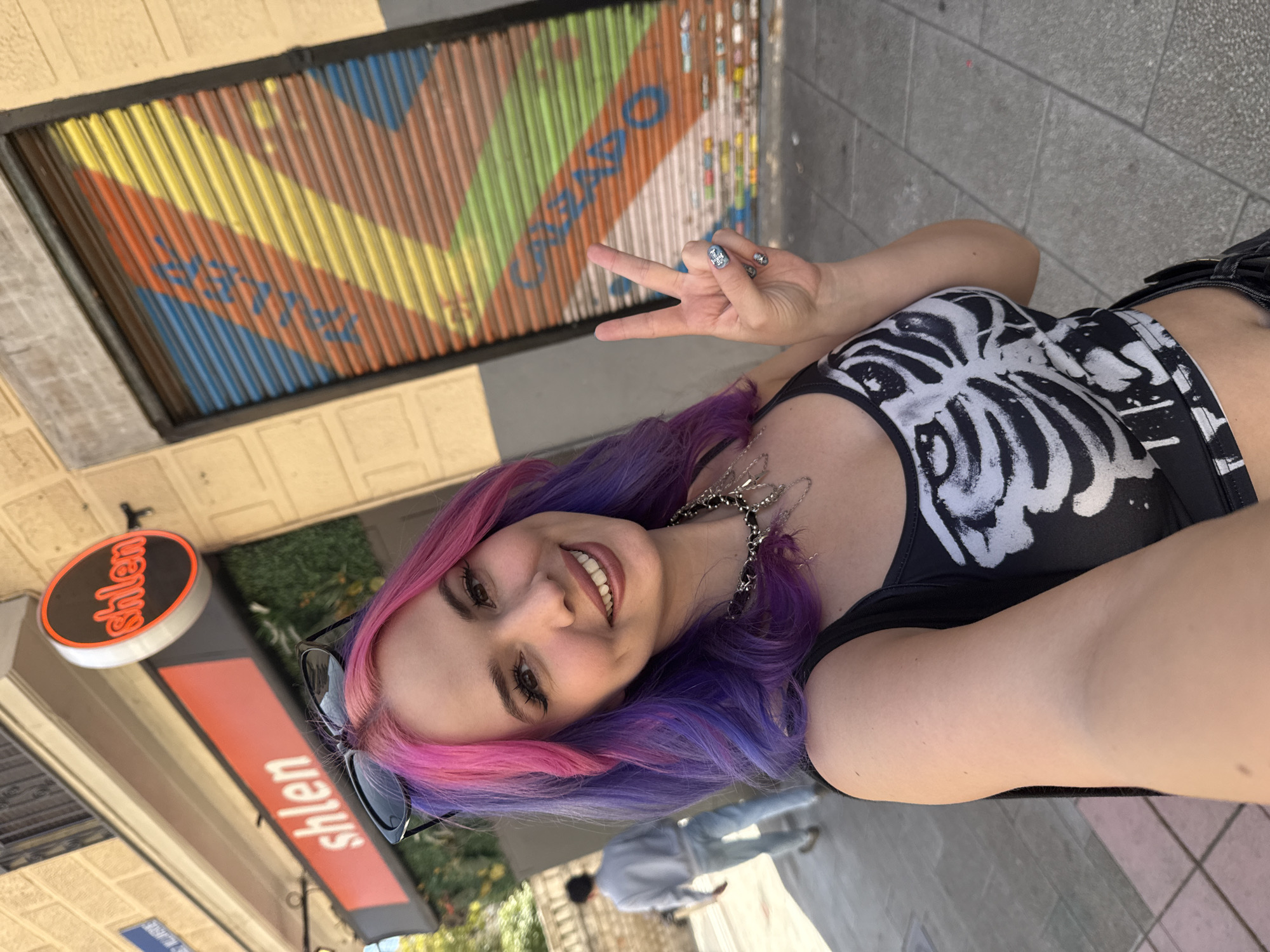 A young woman with pink and purple hair poses for a selfie on a sunny day in Madrid, Spain. She is smiling and holding up a peace sign. She is wearing a black tank top with a white skeleton ribcage print and a silver chain necklace. Sunglasses are perched on her head. Behind her is a closed storefront with colorful corrugated metal shutters. The word "OWEO" is faintly visible on the shutters. An "shlen" sign is visible in the lower-left corner, marking a local business. A person wearing light blue jeans and a light-colored jacket walks behind her. The background suggests a street scene with paving stones.