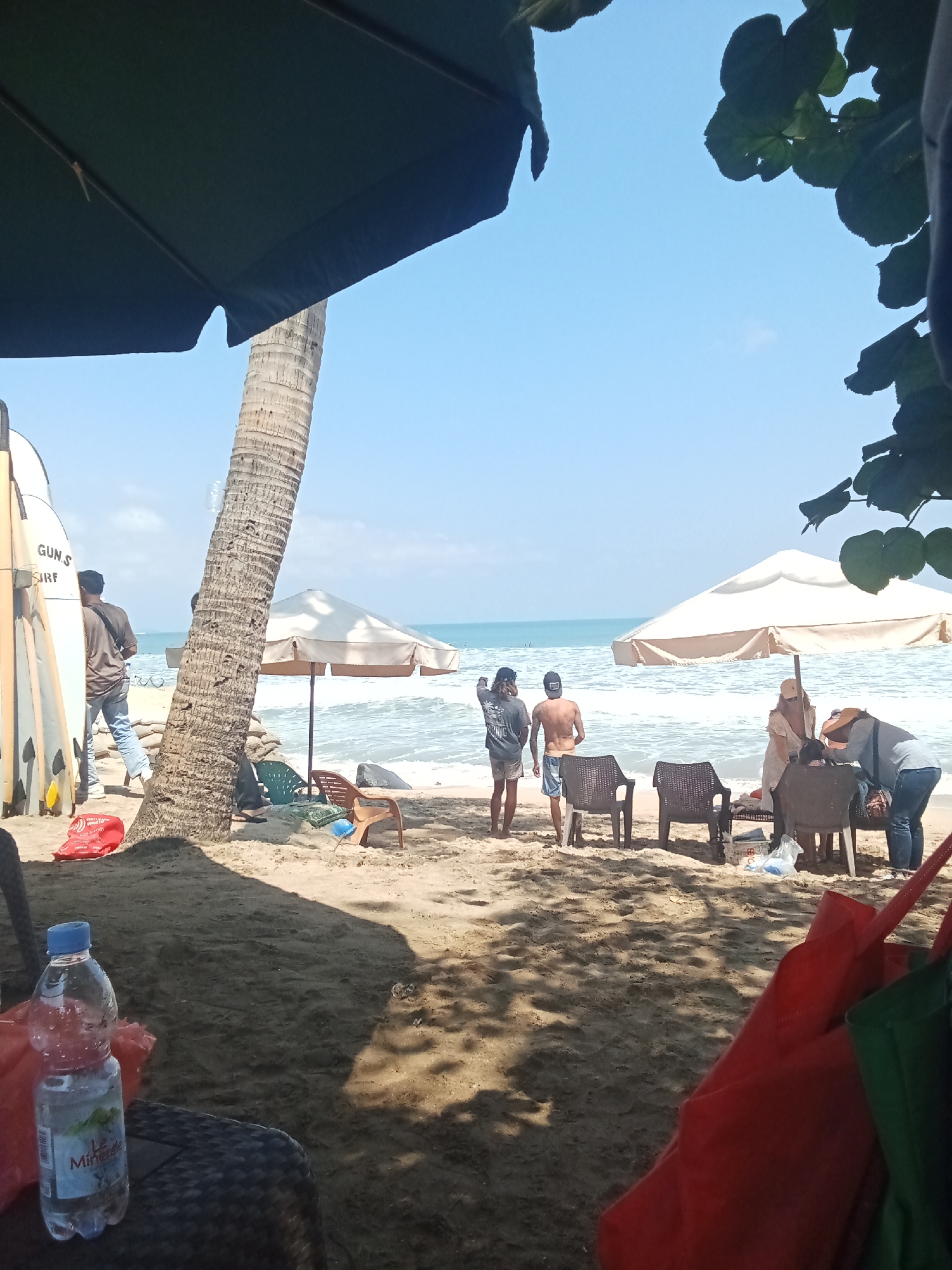 The image captures a beachfront scene in Kuta, Indonesia, characterized by a sandy shore, ocean waves, and a clear blue sky. In the foreground, a plastic water bottle is prominently placed on a woven surface, with red and green bags visible to the right, partially obscuring the view of the sand. A palm tree stands to the left of the frame, its trunk textured and weathered.
Further back, several people are gathered near the water's edge. Two individuals, a man and a woman, stand facing the ocean. A man in a t-shirt and jeans stands near a stack of surfboards on the left side of the image. Two other individuals, a shirtless man and a person wearing a cap, are positioned near the center. Closer to the water, a group of people are interacting under a large, light-colored umbrella, with chairs arranged around them. The ocean is active with visible waves, and a few distant figures can be seen in the water. The overall atmosphere suggests a relaxed, recreational day at the beach.