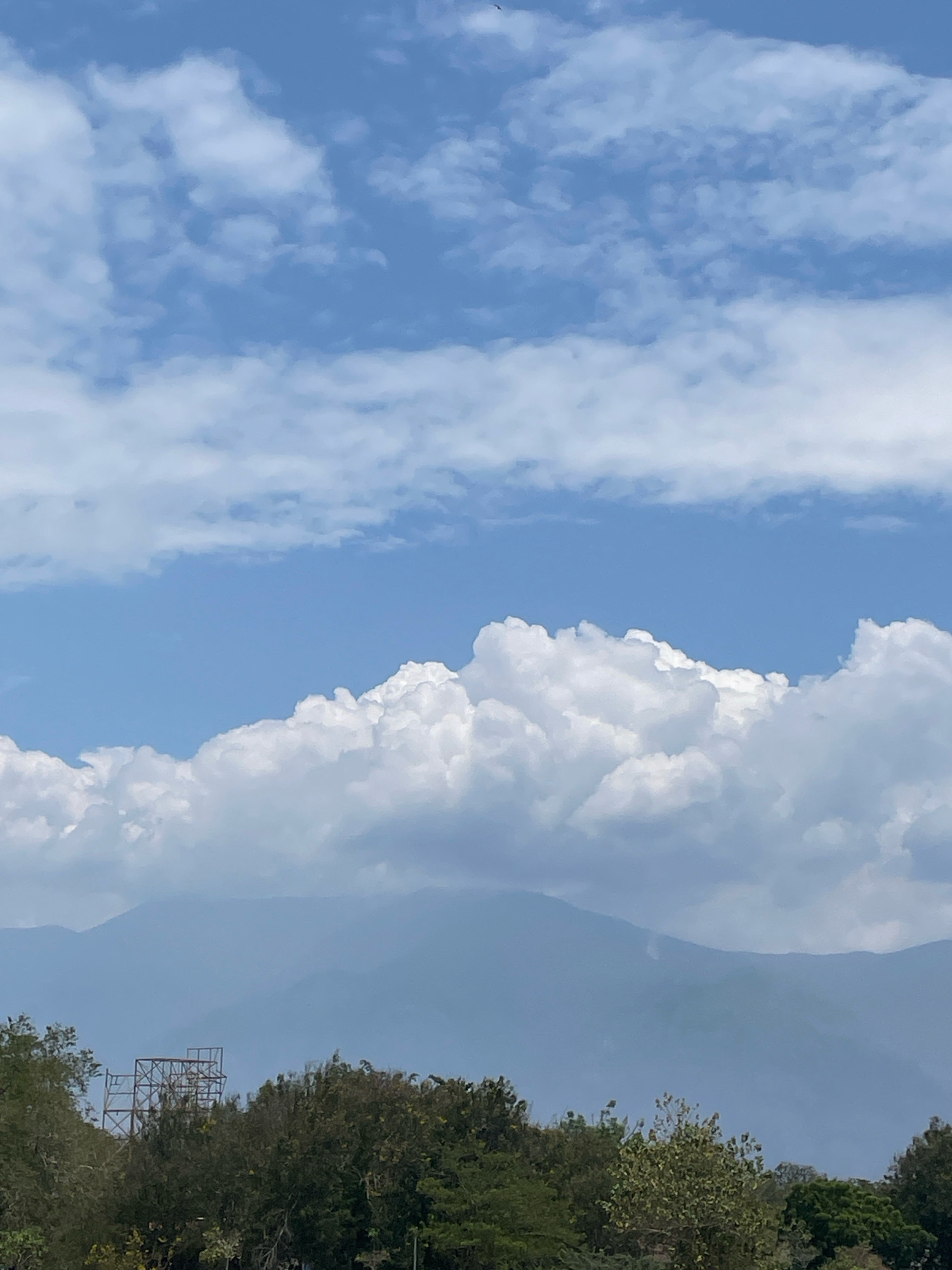 The image captures a wide sky filled with a mix of scattered clouds and larger cumulus formations. Below the sky, a range of hazy blue mountains is visible. In the foreground, a line of green trees stretches across the frame, with a skeletal metal structure, possibly a radio tower or scaffolding, standing amidst the foliage. The scene appears to be an outdoor landscape, with no people or specific city elements of Iradatpuri, Indonesia, identifiable.
